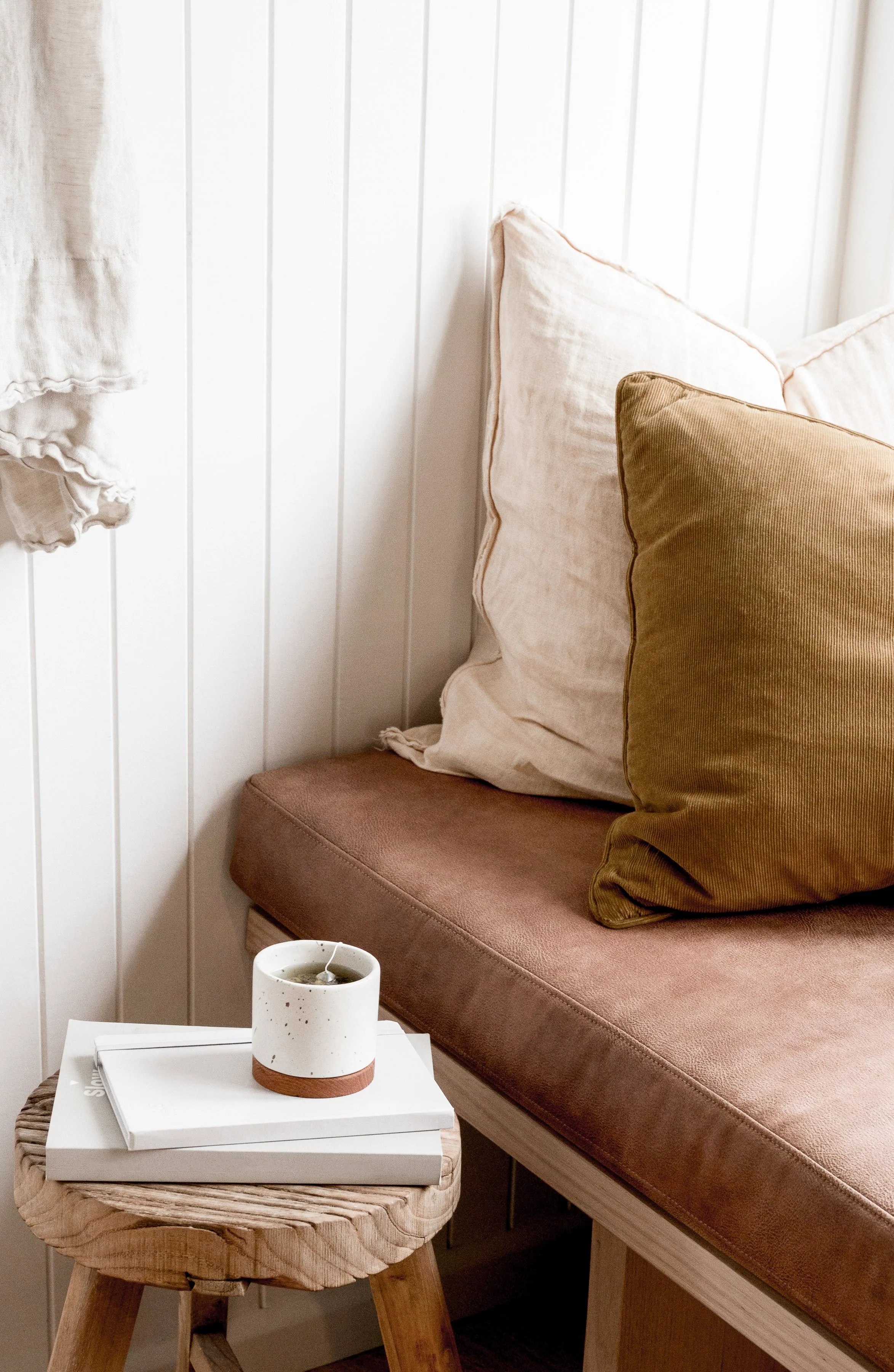 Close-up of a cozy corner with a pink cushioned bench, large beige and brown pillows, a small wooden stool holding a white book and a speckled white ceramic mug, next to white paneled wall.