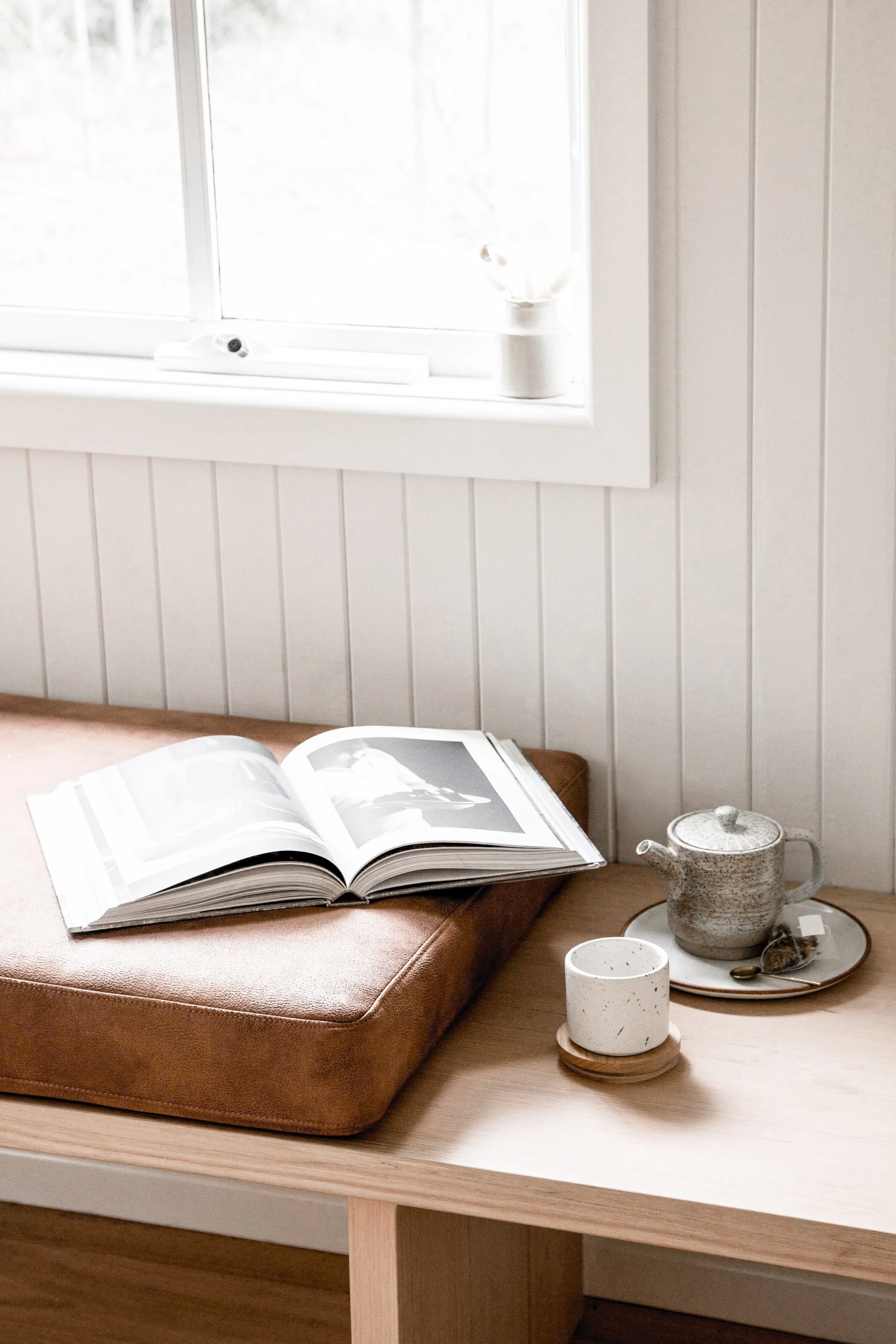 An open book resting on a brown cushion on a wooden table near a window with white frame. Next to the book, there is a ceramic teapot on a tray, and a white cup on a small coaster. The scene is lit with natural light.