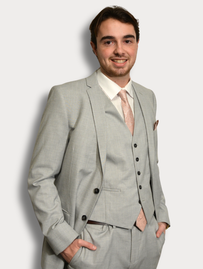 A younger man with short hair, dressed in a grey suit and pink tie, standing against a plain white background.