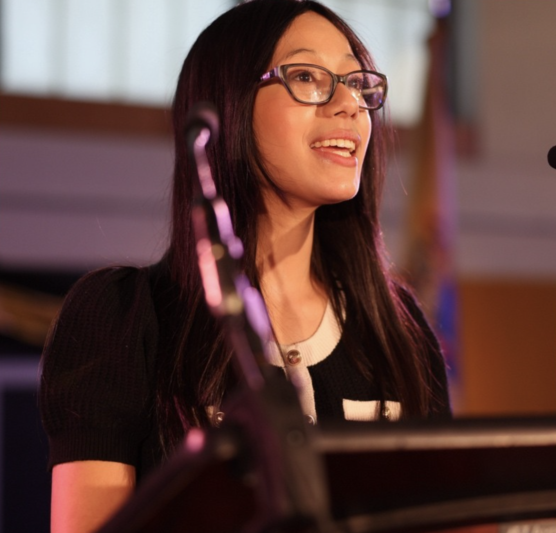 A woman with long dark hair, wearing glasses, smiling and talking, likely performing or speaking at a public event.