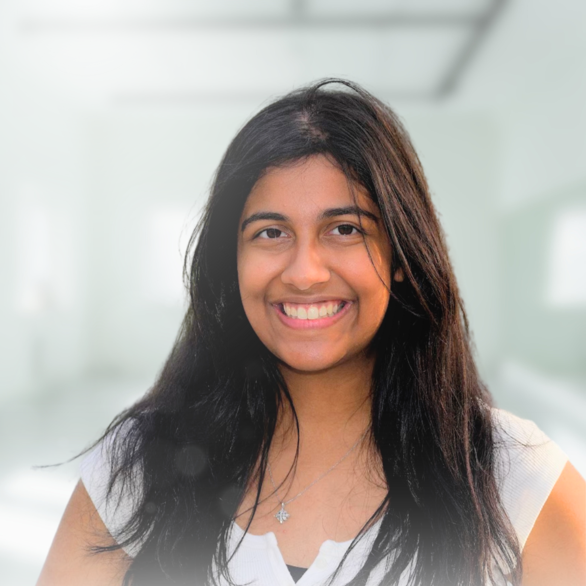 A young woman with long dark hair smiling, wearing a white top and a necklace, with a blurred light-colored background.