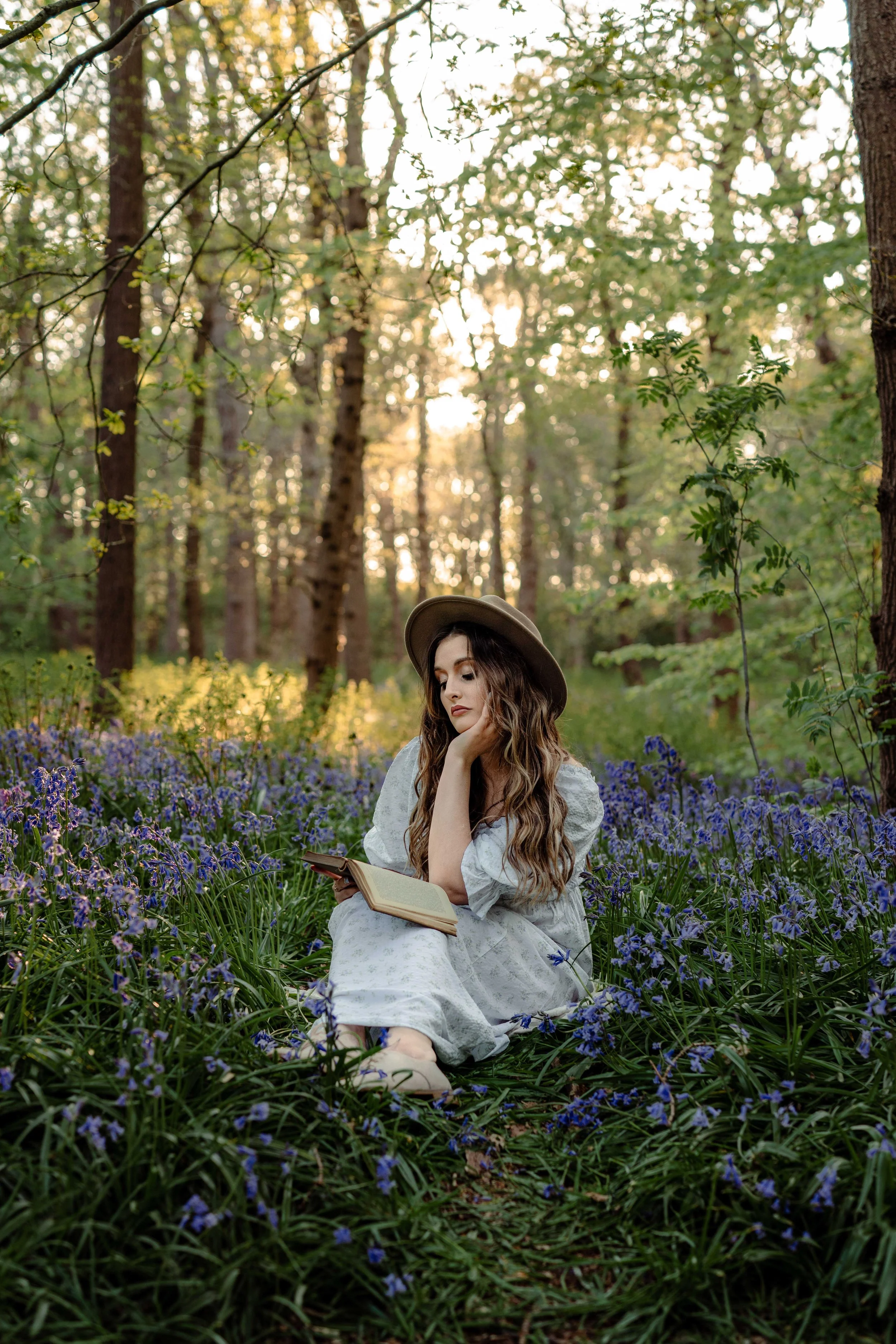 A young woman with long wavy hair, wearing a white dress and a brown hat, sitting in a forested area surrounded by purple wildflowers, reading a book during sunset. Romantic portrait photoshoot in spring in The Hague.