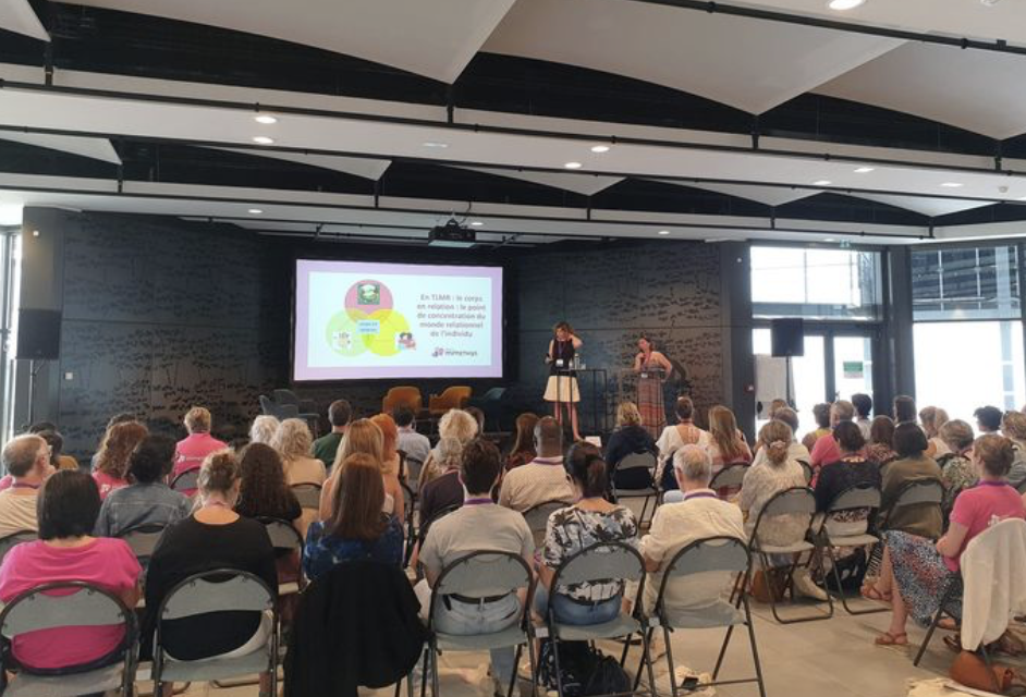 Une conférence ou un séminaire avec des participants assis face à une scène où deux personnes parlent devant un grand écran. La salle est moderne avec de grandes fenêtres et un plafond à plusieurs niveaux.