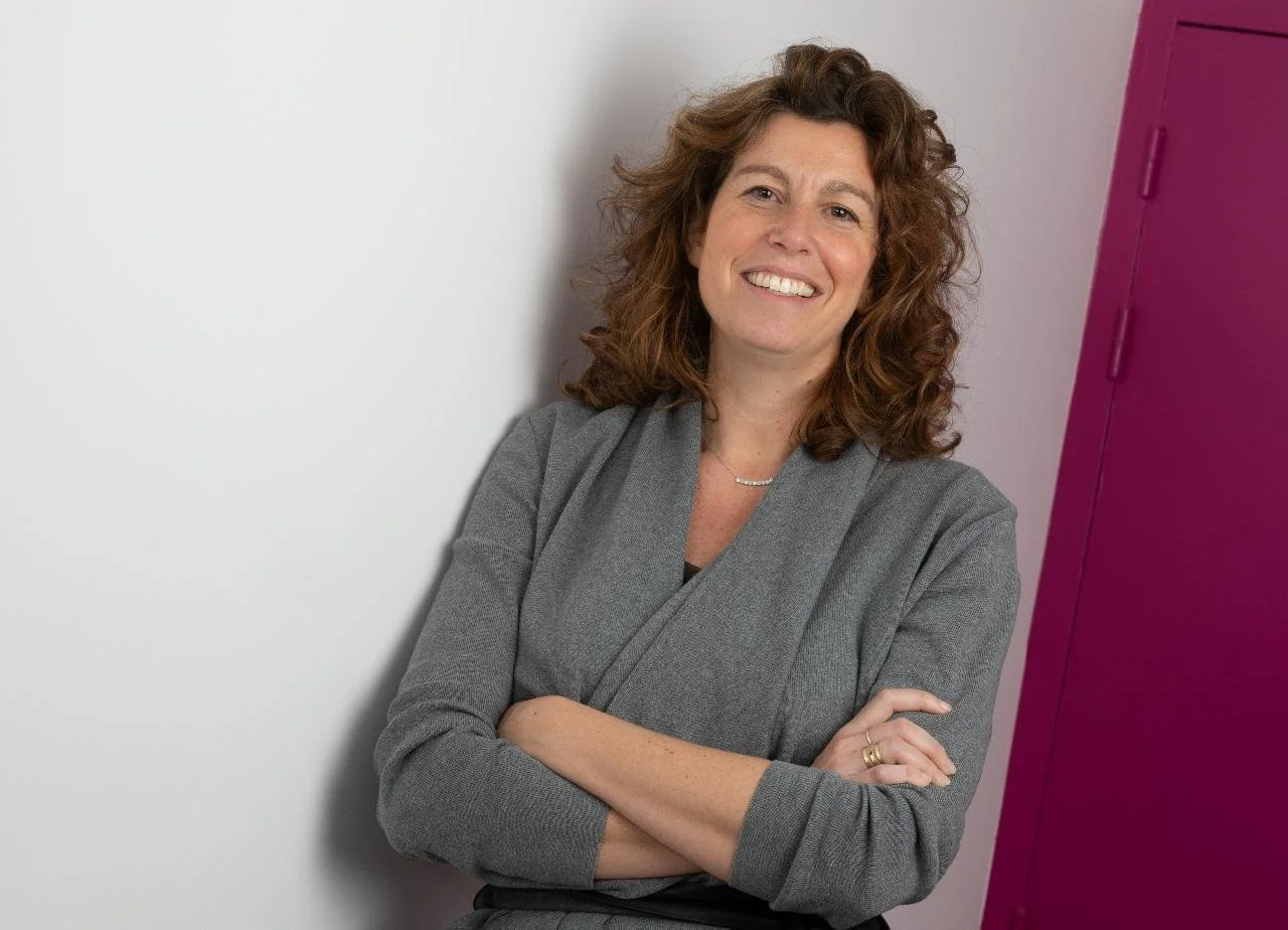 Une femme souriante avec des cheveux bouclés, portant un blazer gris, debout contre un mur blanc et une armoire violette.