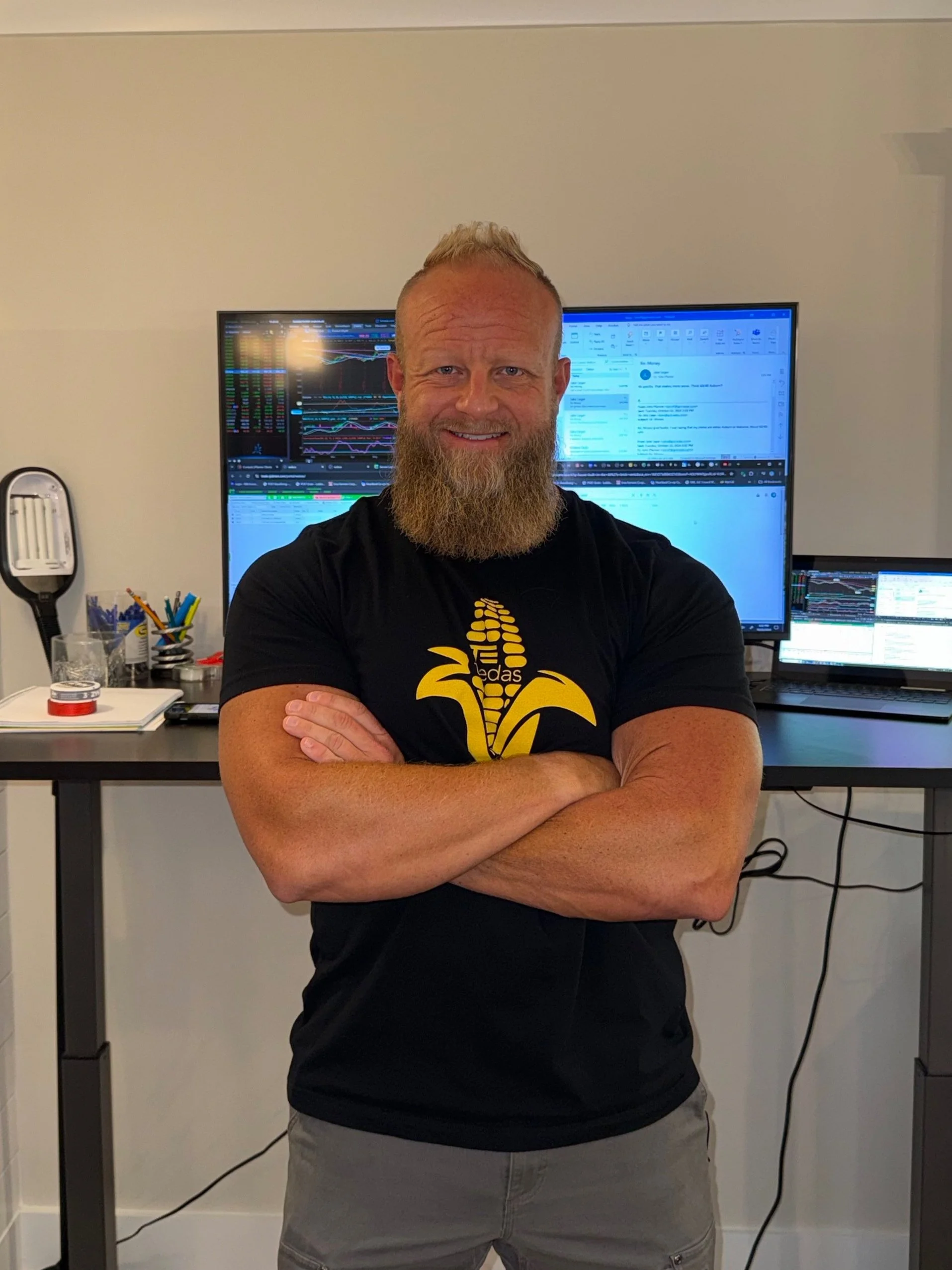 A man with a beard, wearing a black T-shirt with a yellow design, standing with arms crossed in front of a desk with multiple monitors displaying financial data and graphs.