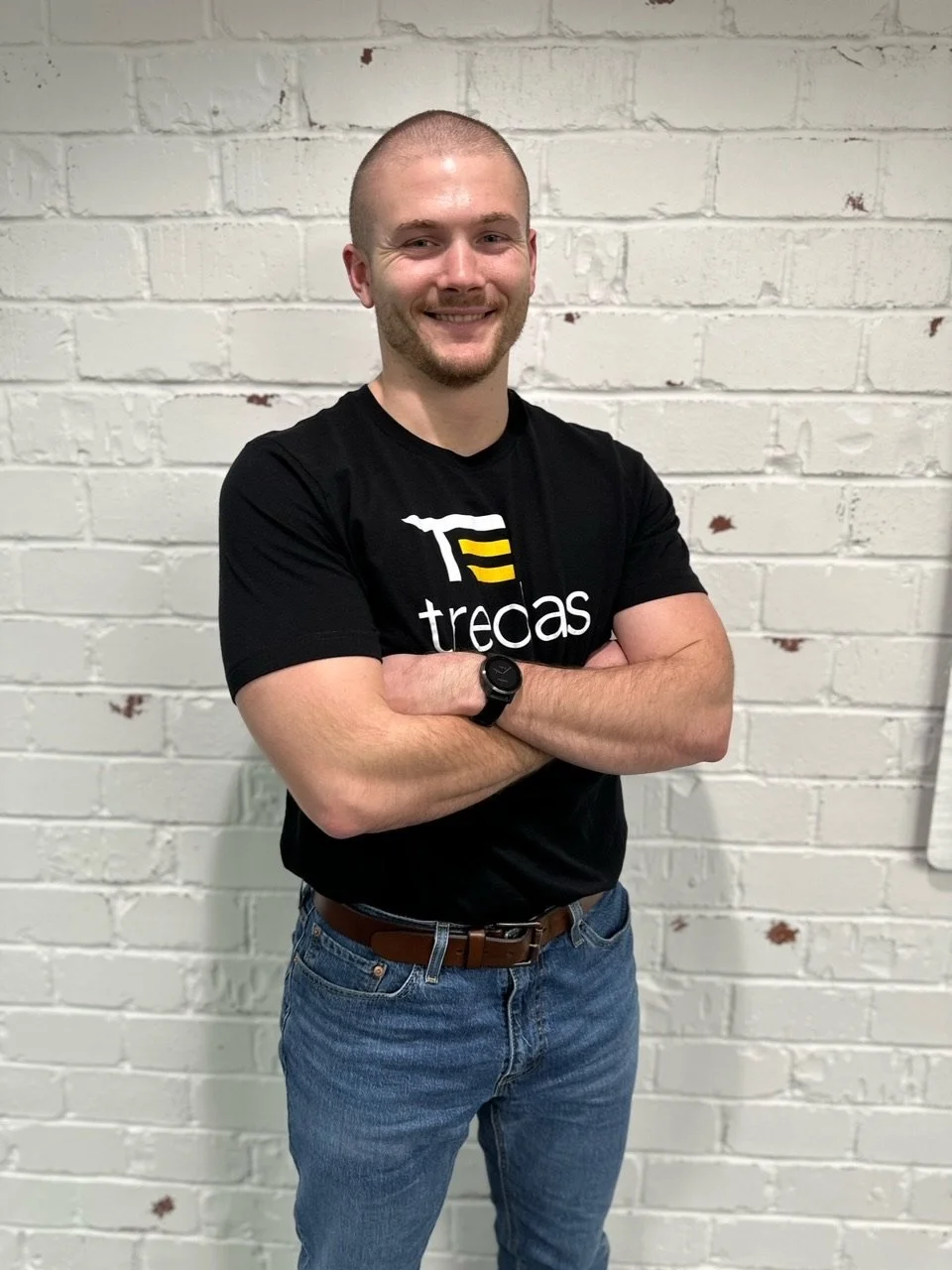 A smiling man with a shaved head and beard, wearing a black Trecas t-shirt, blue jeans, and a black watch, standing with arms crossed in front of a white brick wall.