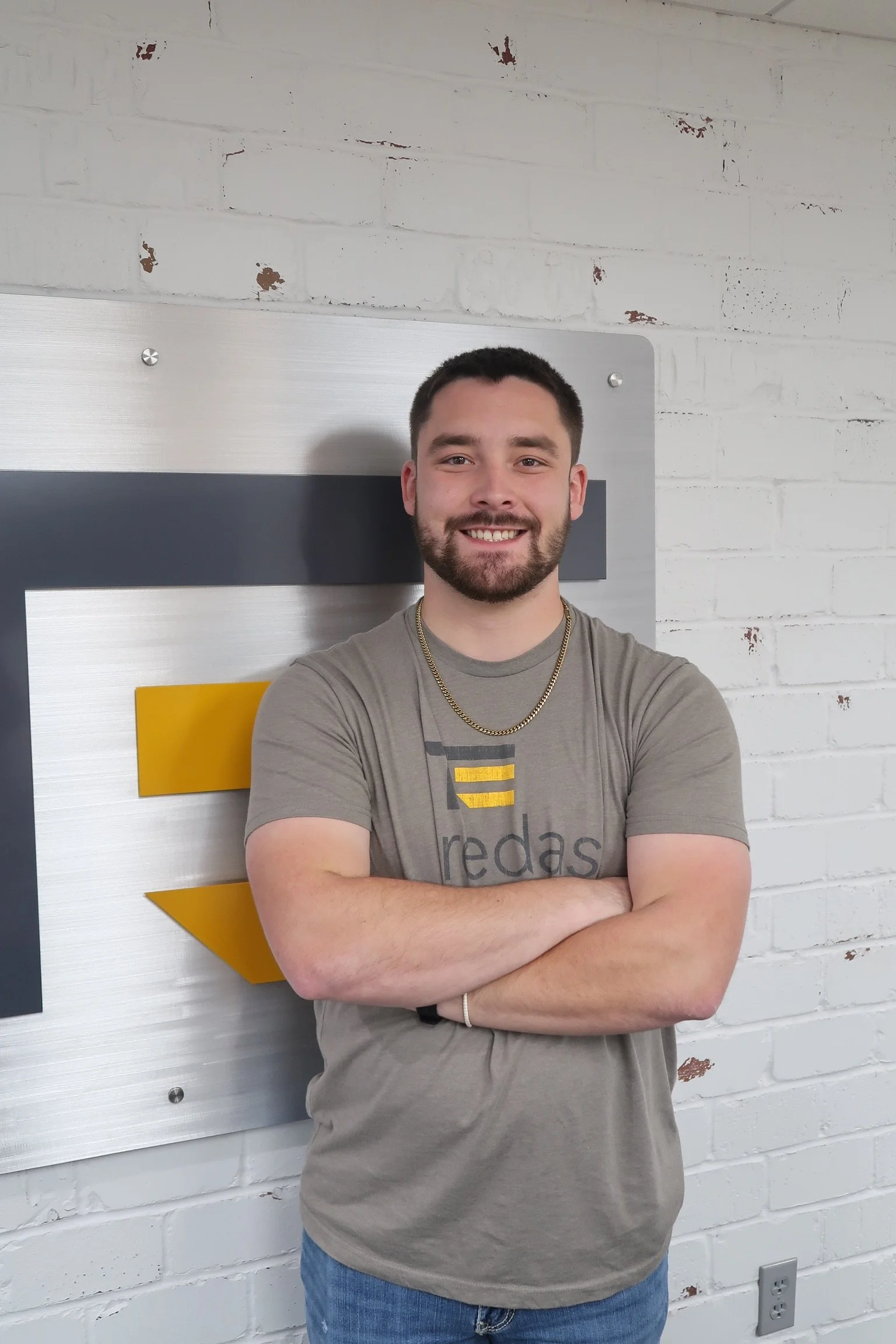 A young man with short dark hair and beard, smiling with arms crossed, standing in front of a white brick wall with a modern sign.