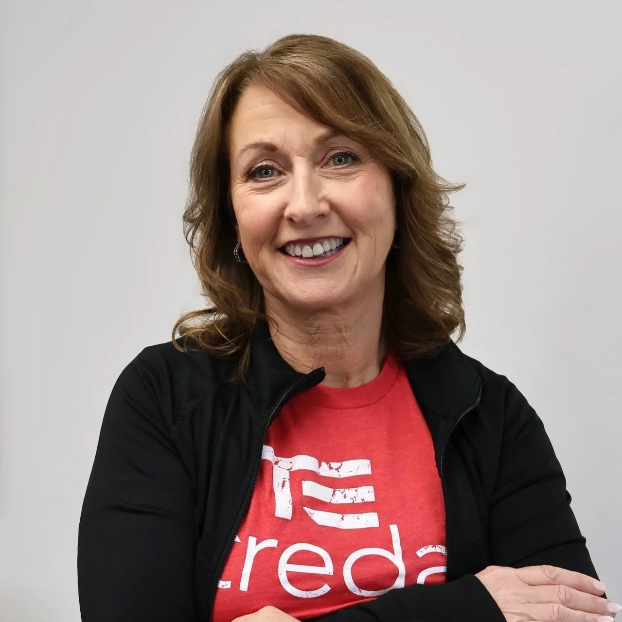 A smiling middle-aged woman with shoulder-length brown hair, wearing a black jacket over a red T-shirt with white text and a graphic, standing against a plain gray background.