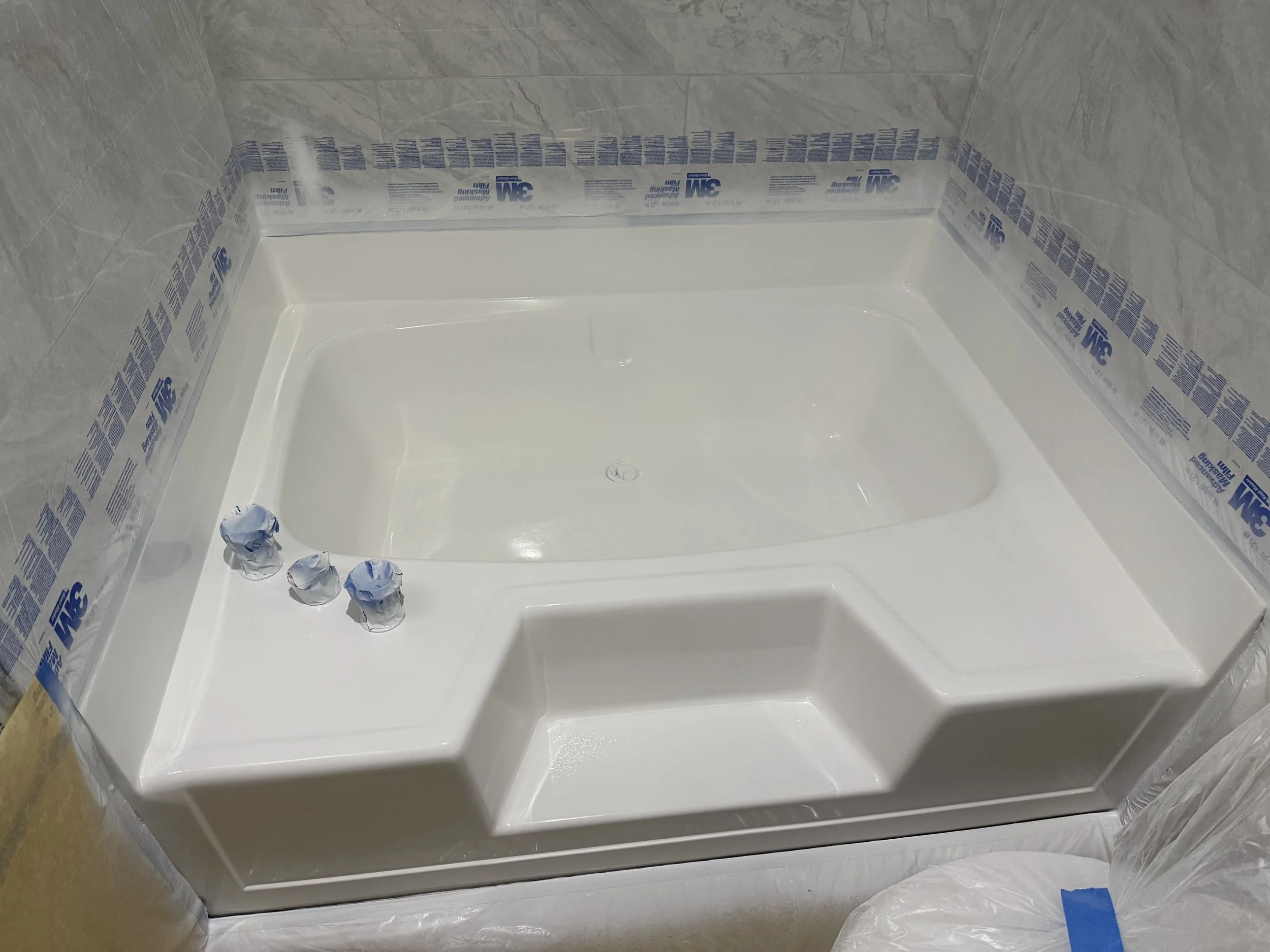 A custom shower with a white acrylic tub, gray tiled walls, and three blue protective caps on the drain cover.