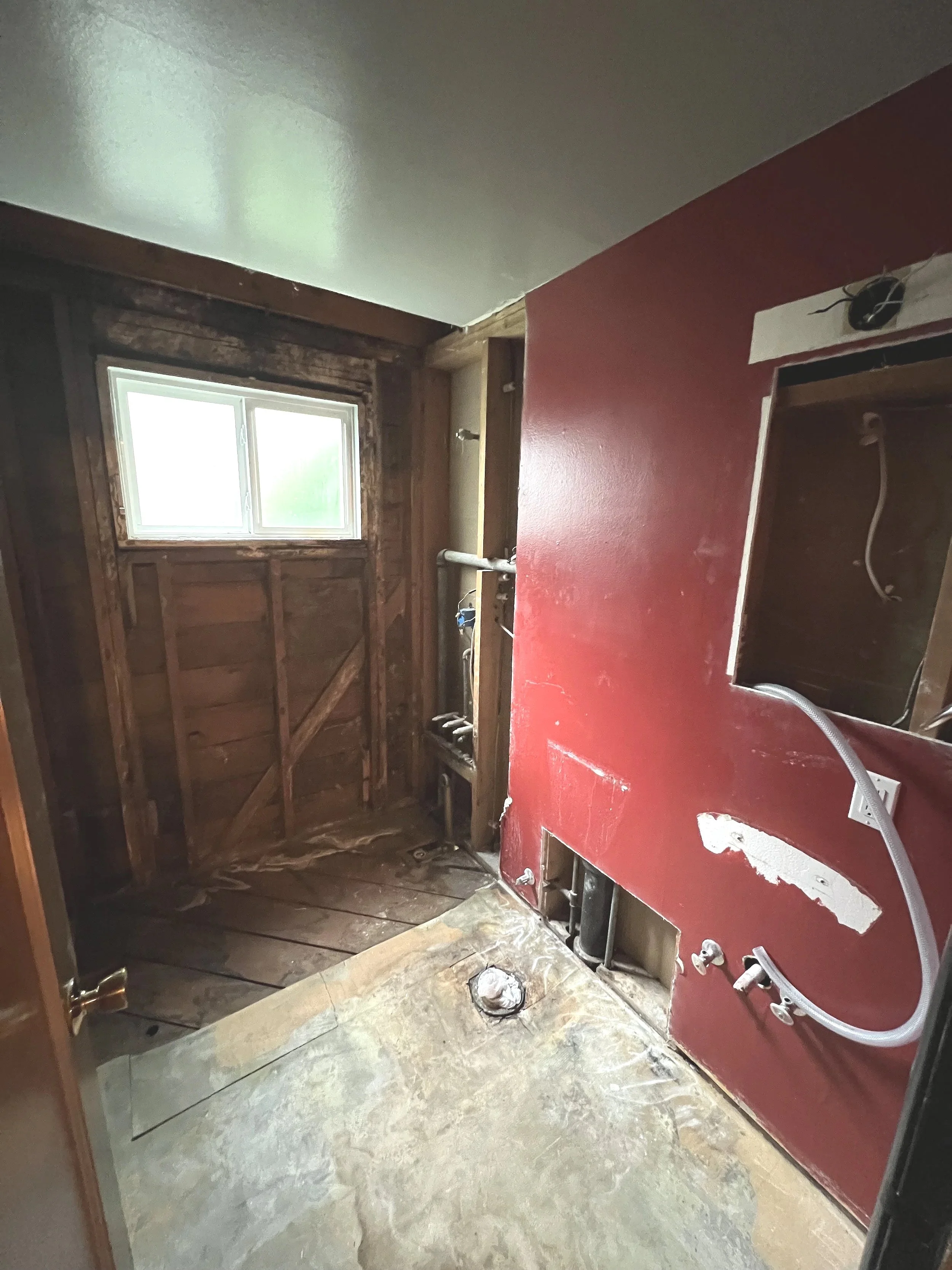 Room under renovation with exposed wood framing, a small window, and plumbing fixtures for a bathroom or laundry area.