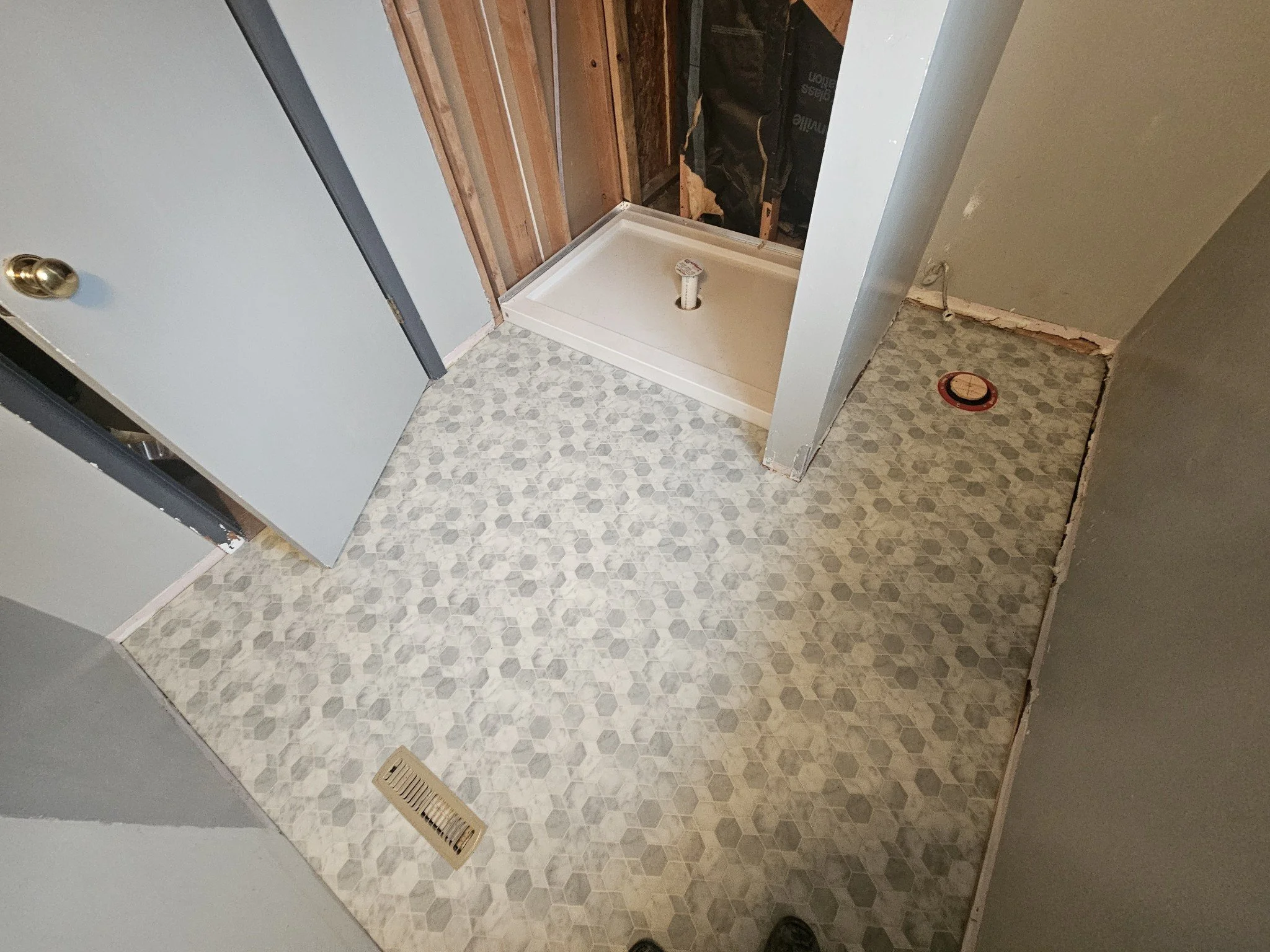 Room with hexagon-patterned vinyl flooring, a vent on the floor, an unfinished shower with a drainage pipe, and a partially installed wall with visible framing and drywall.