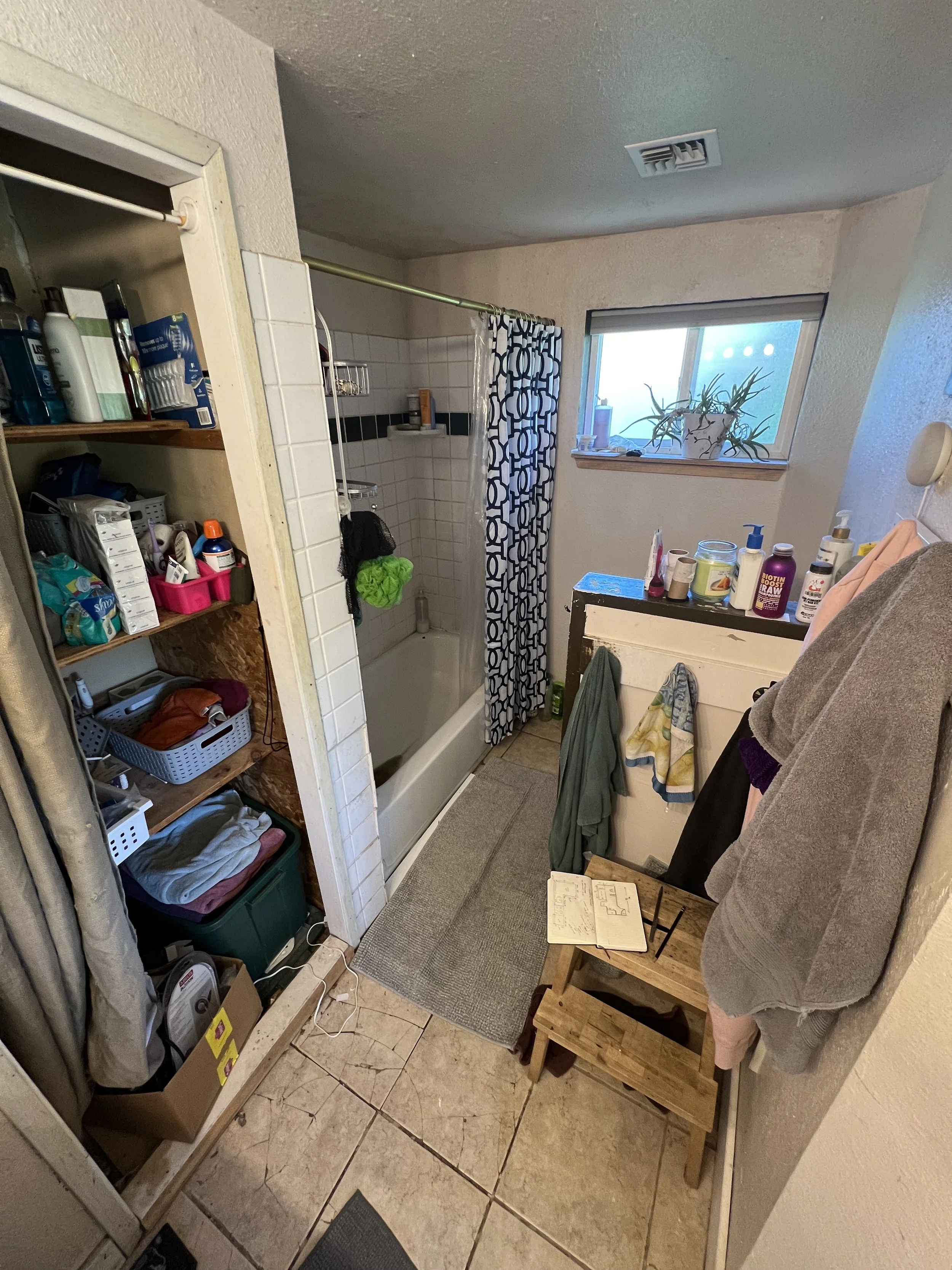 View of a small bathroom with an open shower, a window with a potted plant, a shelf with toiletries, and various scattered items and towels.