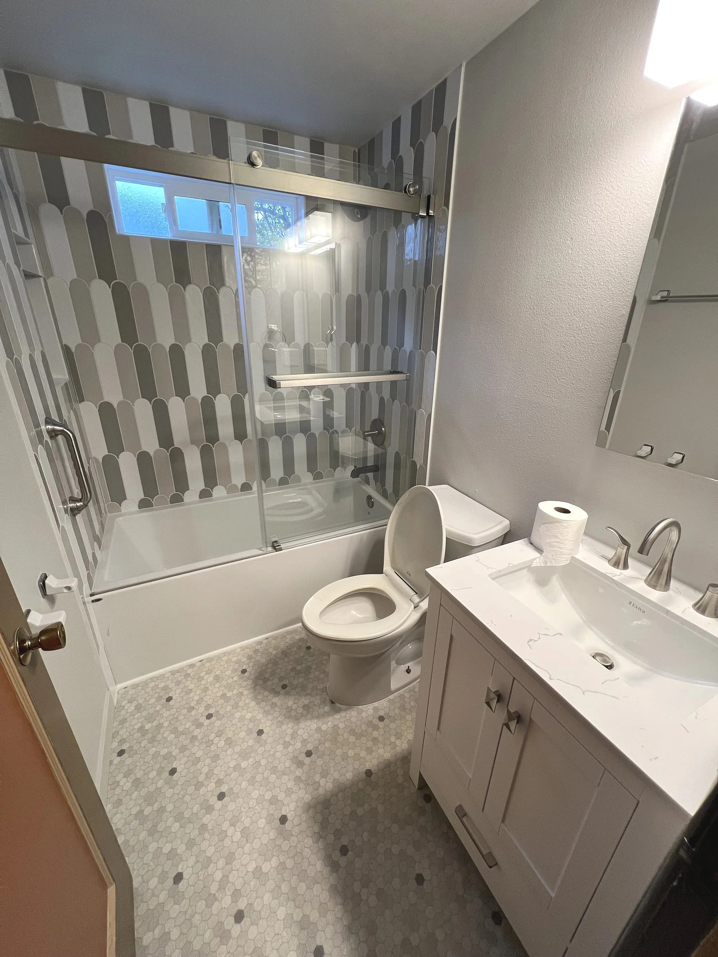 A small bathroom with a white vanity sink, a toilet with the lid up, a bathtub with a glass sliding door, patterned gray and white tiles on the wall in the shower, a small window above the shower, a mirror above the sink, and a roll of toilet paper on the sink.