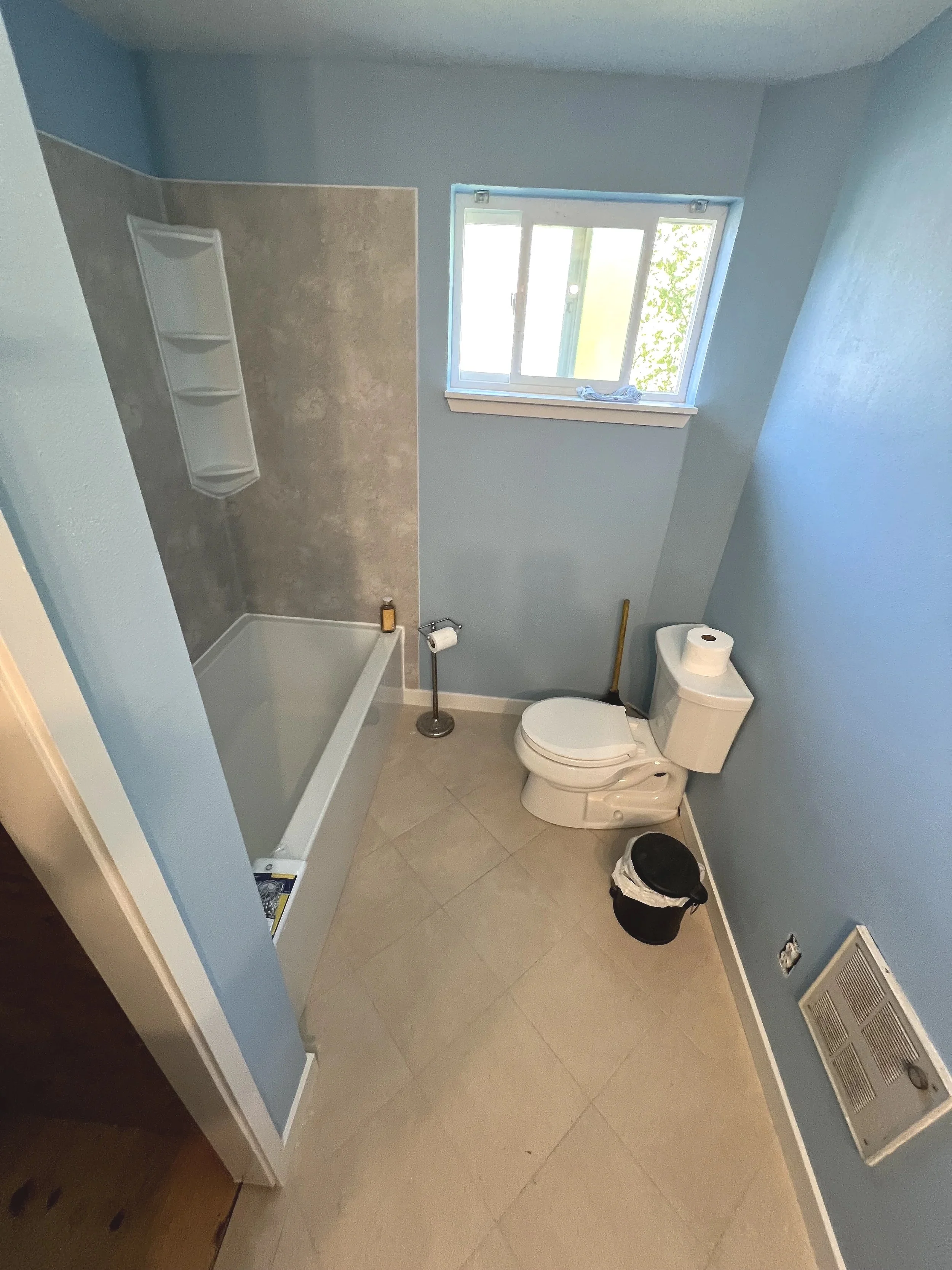 A small, partially renovated bathroom with light blue walls, a small window, a toilet with a roll of toilet paper on top, a small black trash bin, a bathtub, a soap shelf, and a vent on the wall.