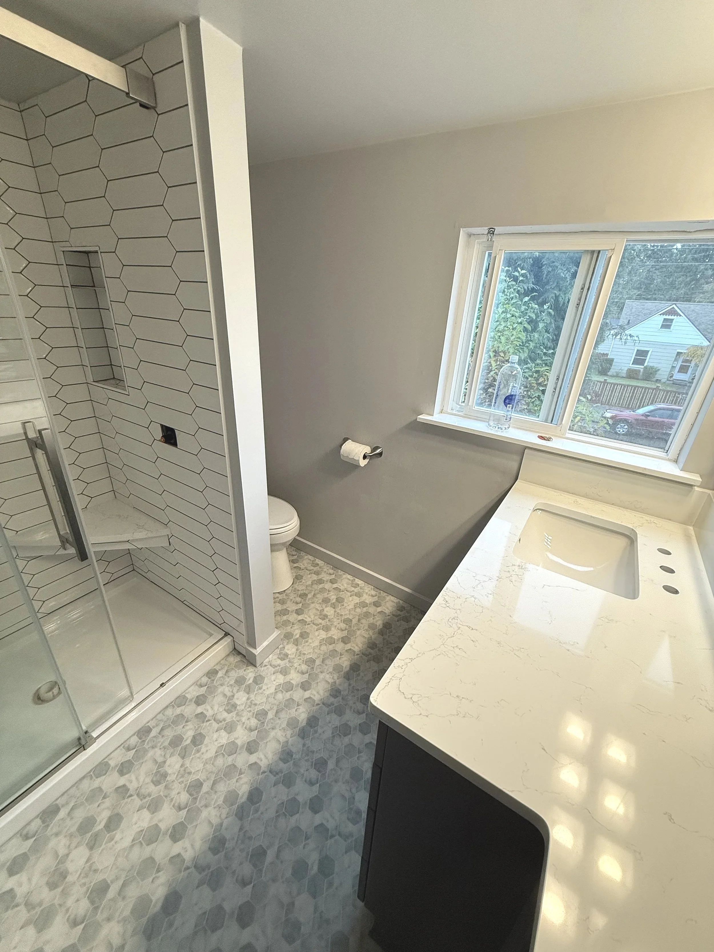 Bathroom with walk-in shower, toilet, and white marble countertop vanity near a window.