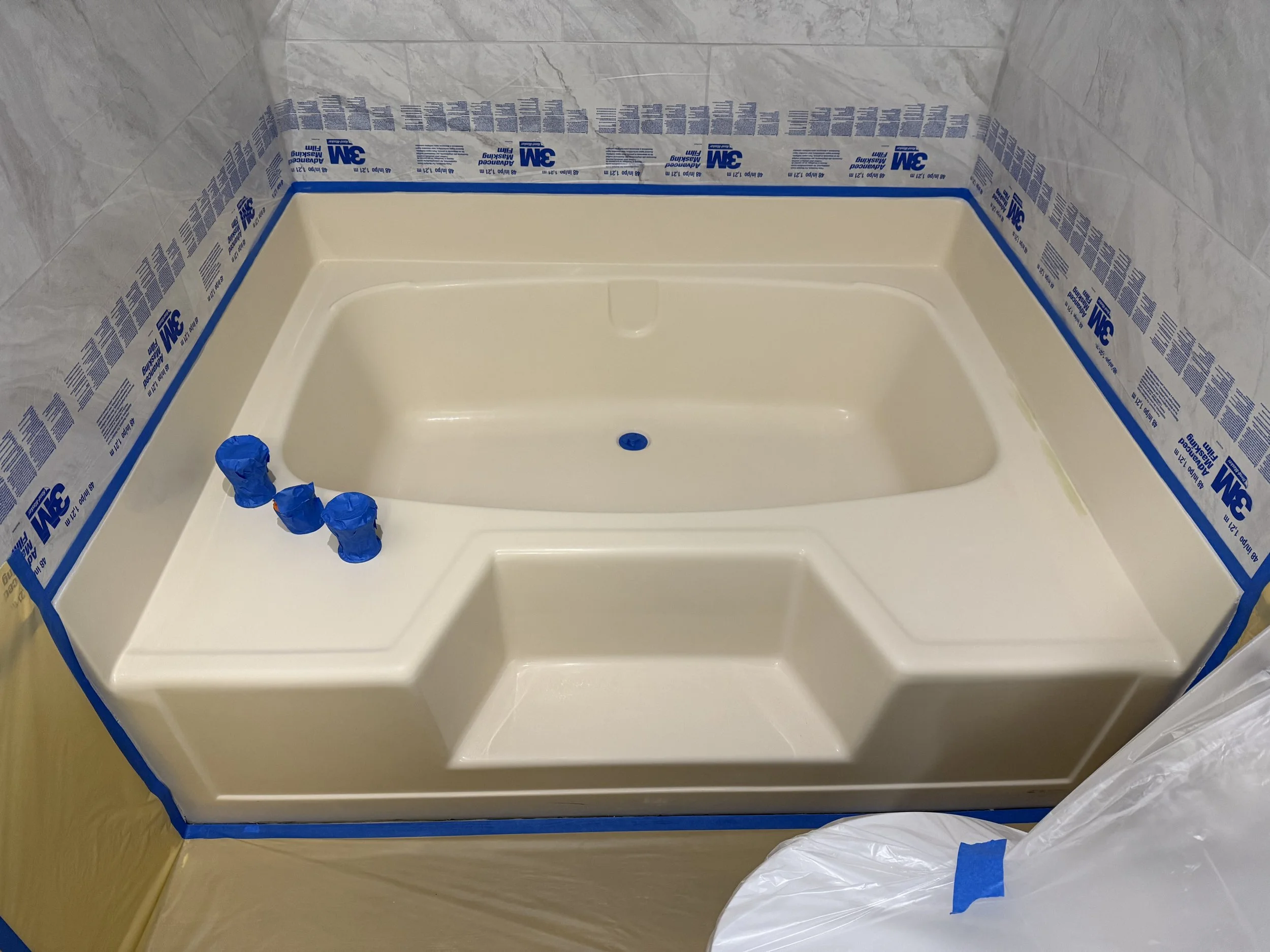 A bathtub under construction with masking tape and blue painter's tape around the edges and on the faucet area in a bathroom with tiled walls.