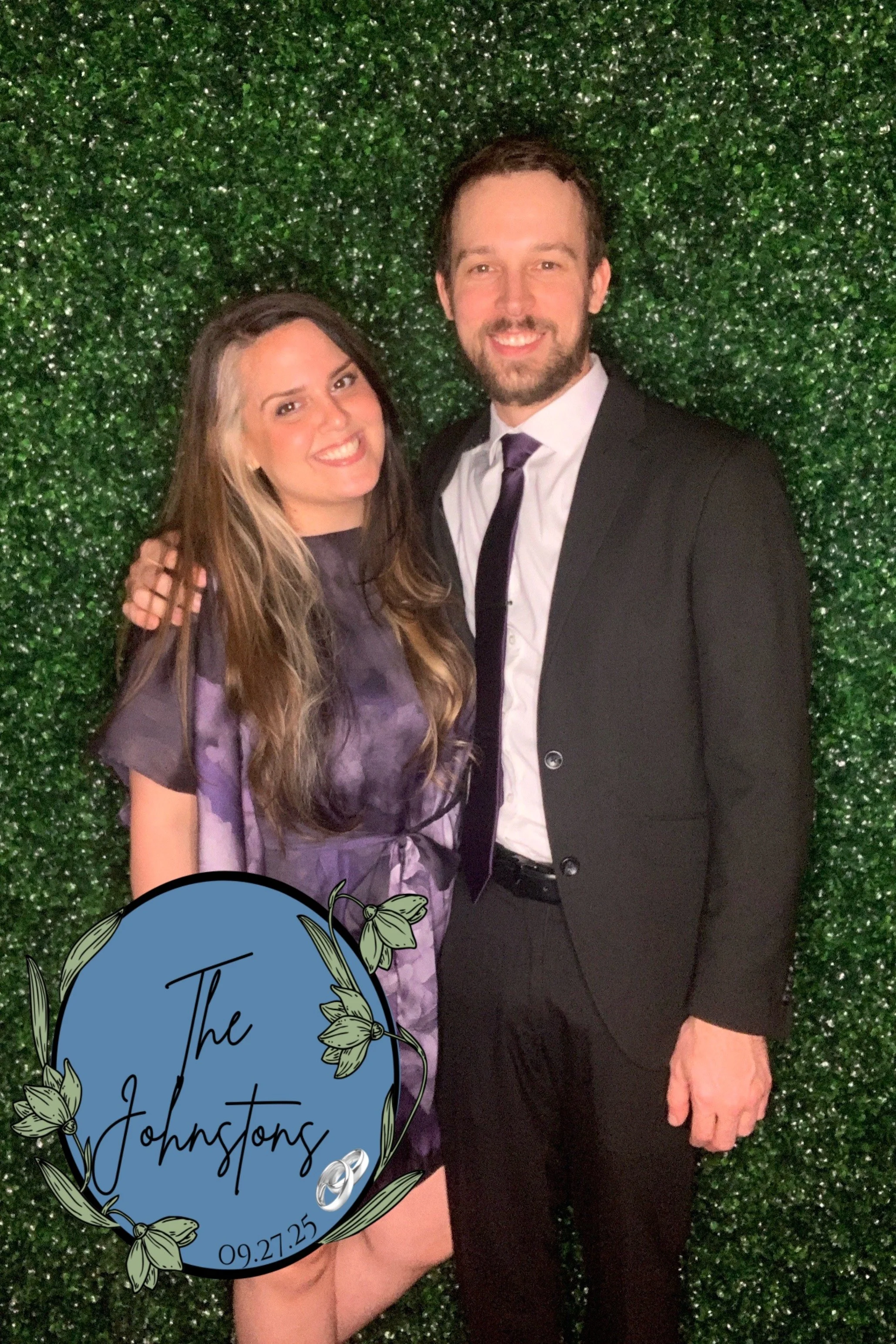 A smiling woman and man posing together in front of a green leafy backdrop at a wedding or special event. The woman has long, wavy hair and is wearing a purple dress. The man has short hair, a beard, and is wearing a black suit with a white shirt and