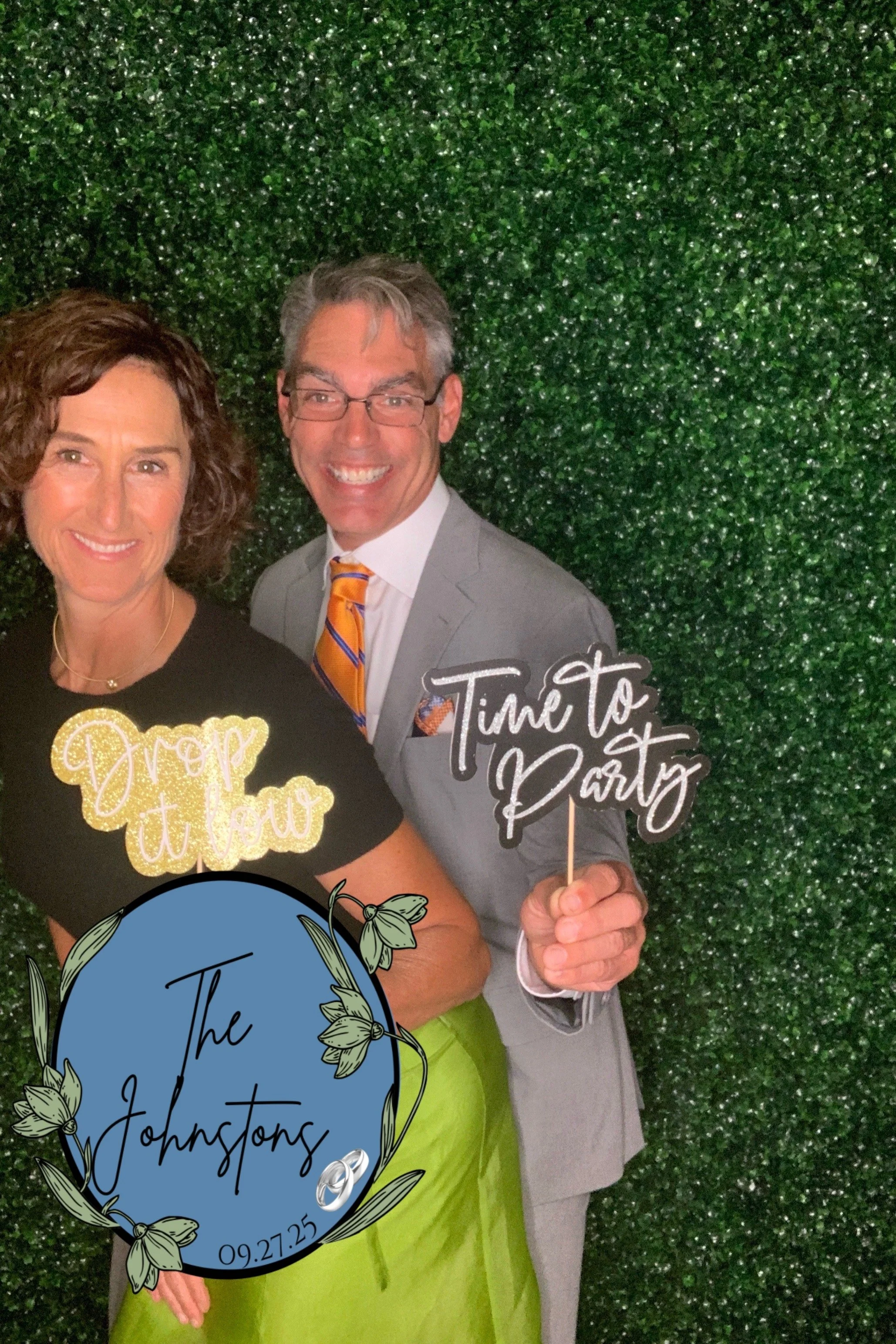 Two people standing in front of a green leafy backdrop, smiling and holding signs that say 'Time to Party' and 'Drop it low,' and a sign with the text 'The Johnstons' and a wedding date, 09.27.25, along with a decorative ring graphic.
