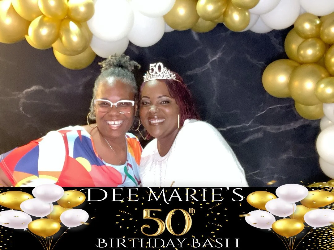Two women smiling at a birthday party, one wearing a tiara that says 50, in front of a balloon decoration with gold and white balloons, for Dee Marie's 50th birthday celebration.