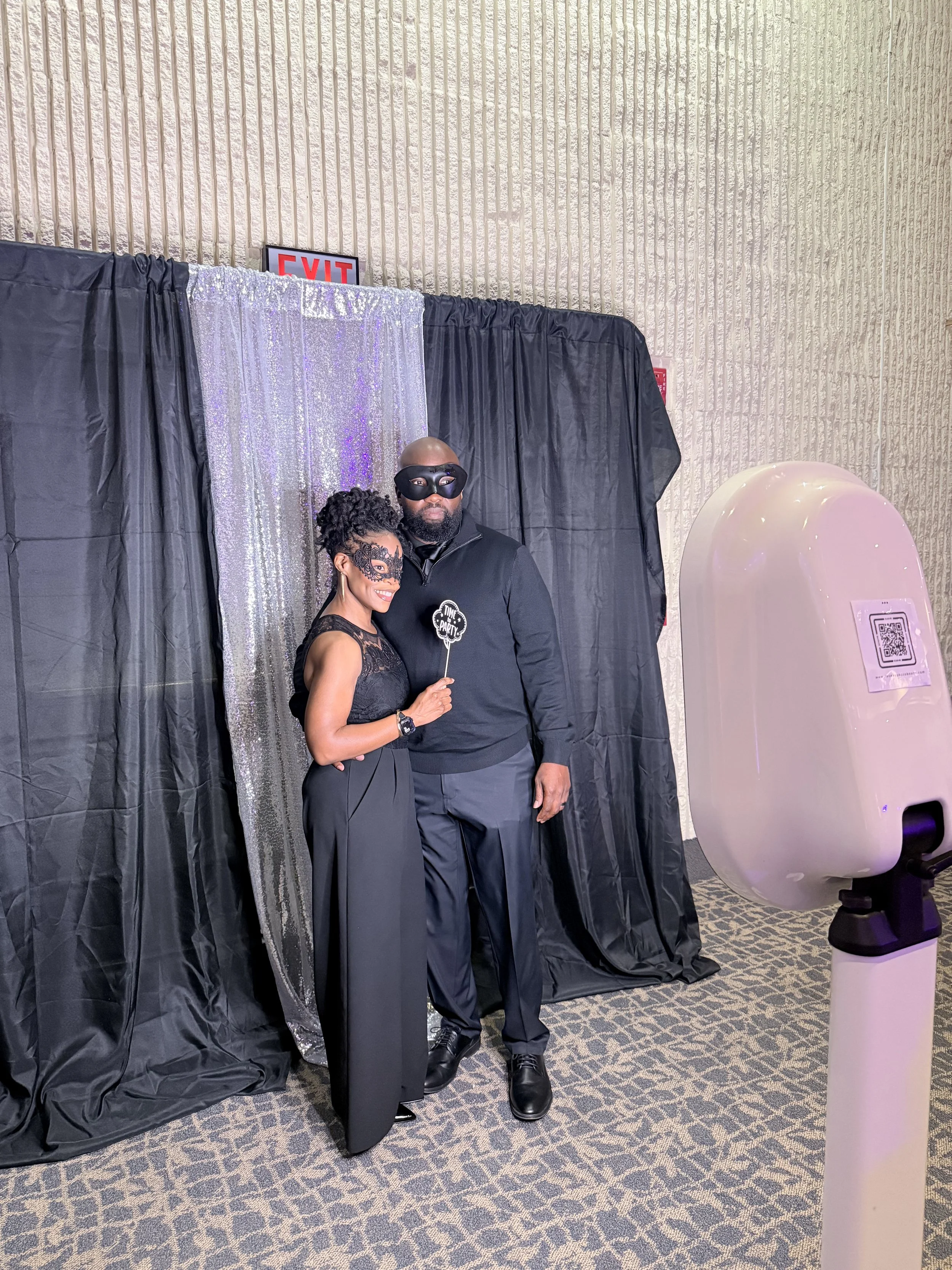 A woman and a man dressed in black posing for a photo at a photo booth with black and glittering silver backdrops, both wearing masks, with the woman holding a small black and white sign that says 'I'm the party'.