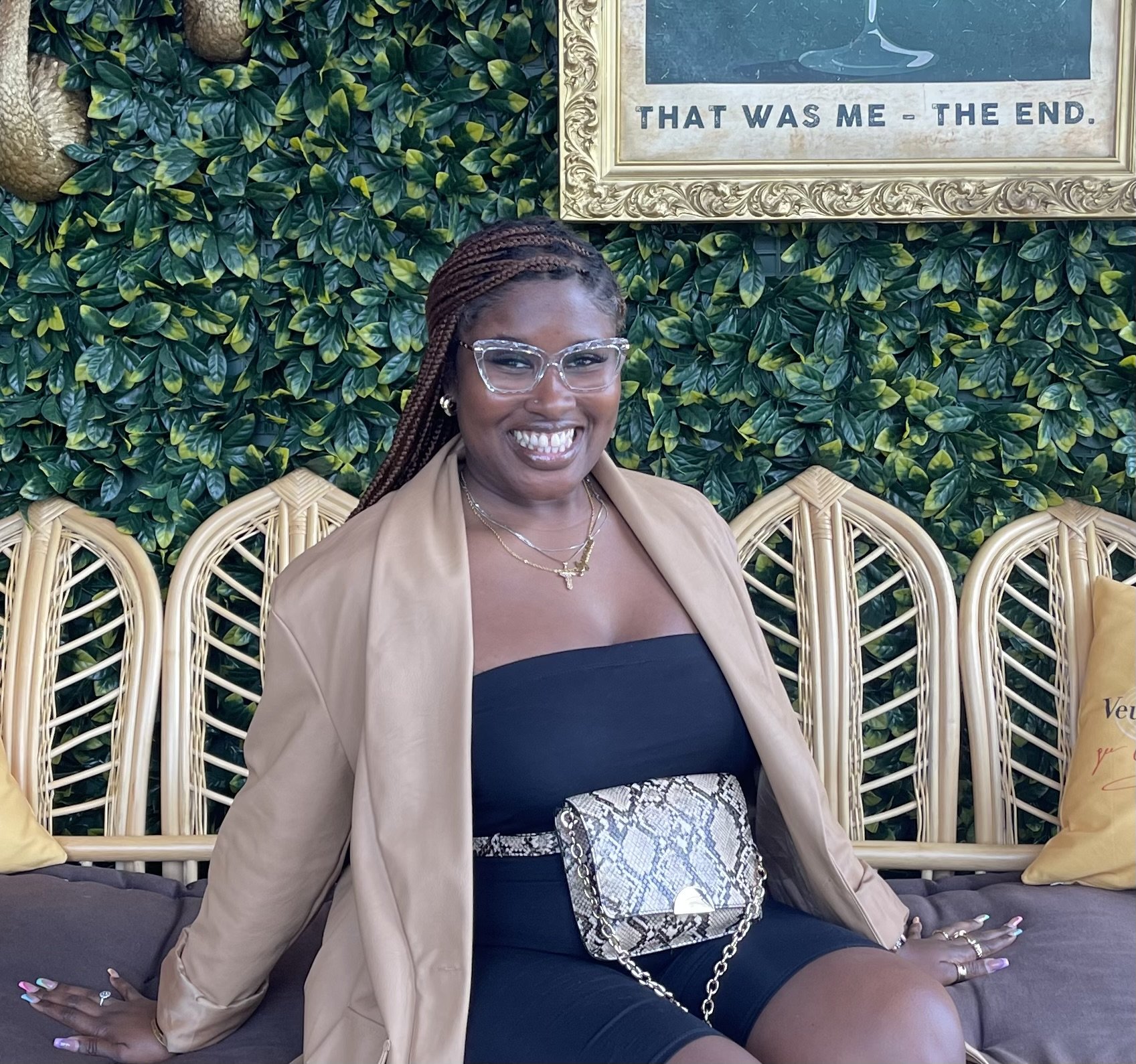 A smiling woman with braided hair and glasses, wearing a beige blazer, black dress, and snakeskin purse, sitting on a rattan bench against a green leafy wall with framed art and yellow pillows.