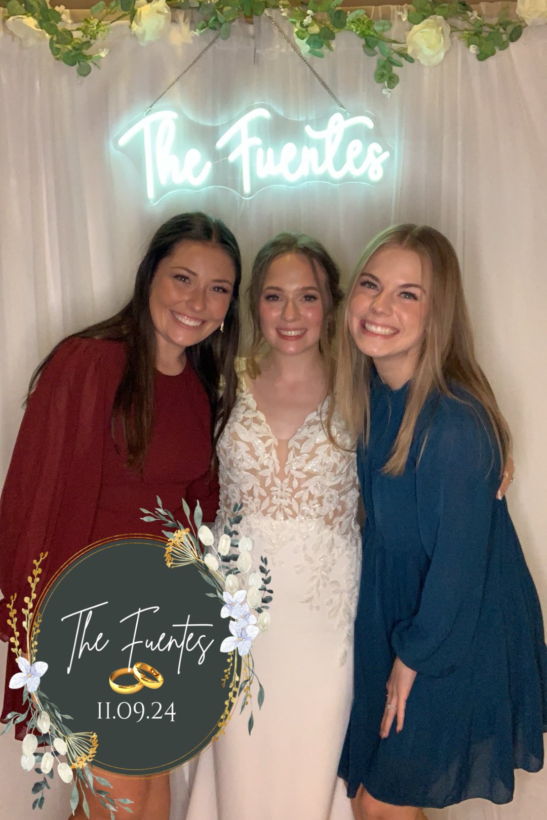 Three women smiling at a wedding. The woman in the middle is wearing a white lace wedding dress, with a neon sign above that reads 'The Fuentes.' The two women on either side are wearing dresses, one in red and the other in blue. There is a graphic o