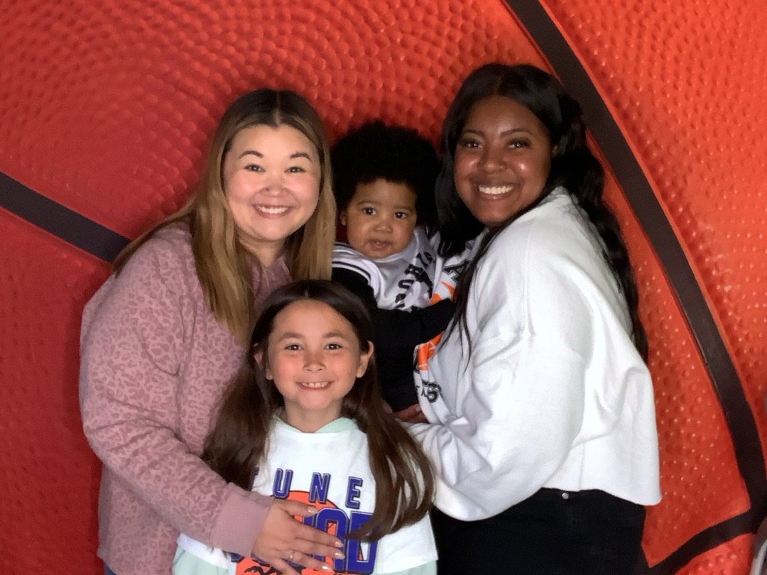 Two women and two children smiling in front of a red, textured background, with one woman holding a young child and the other holding a girl.
