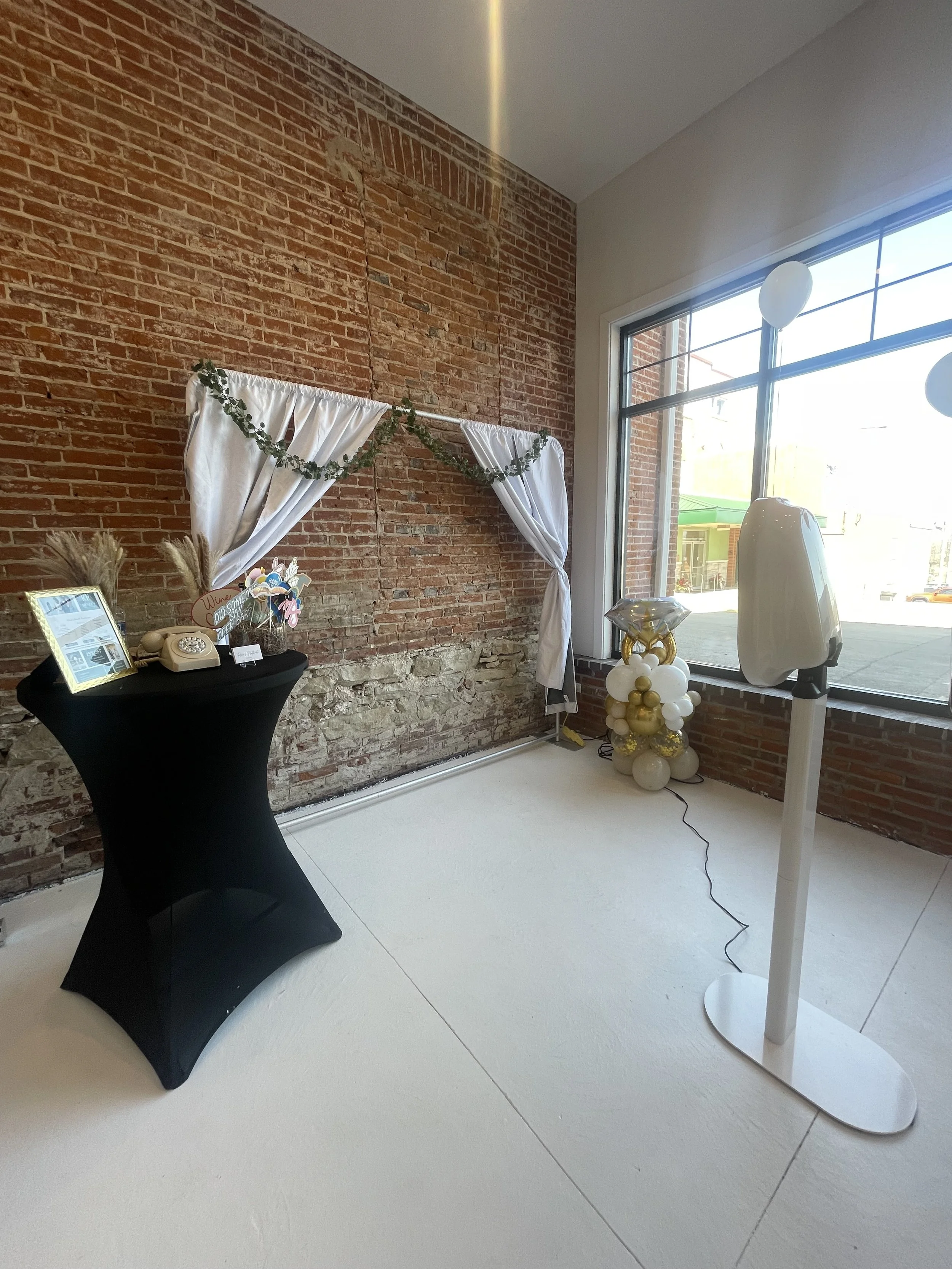 Decorated photo booth area with white curtains, balloon bouquet, and a table with photo props inside a room with exposed brick walls and large window.