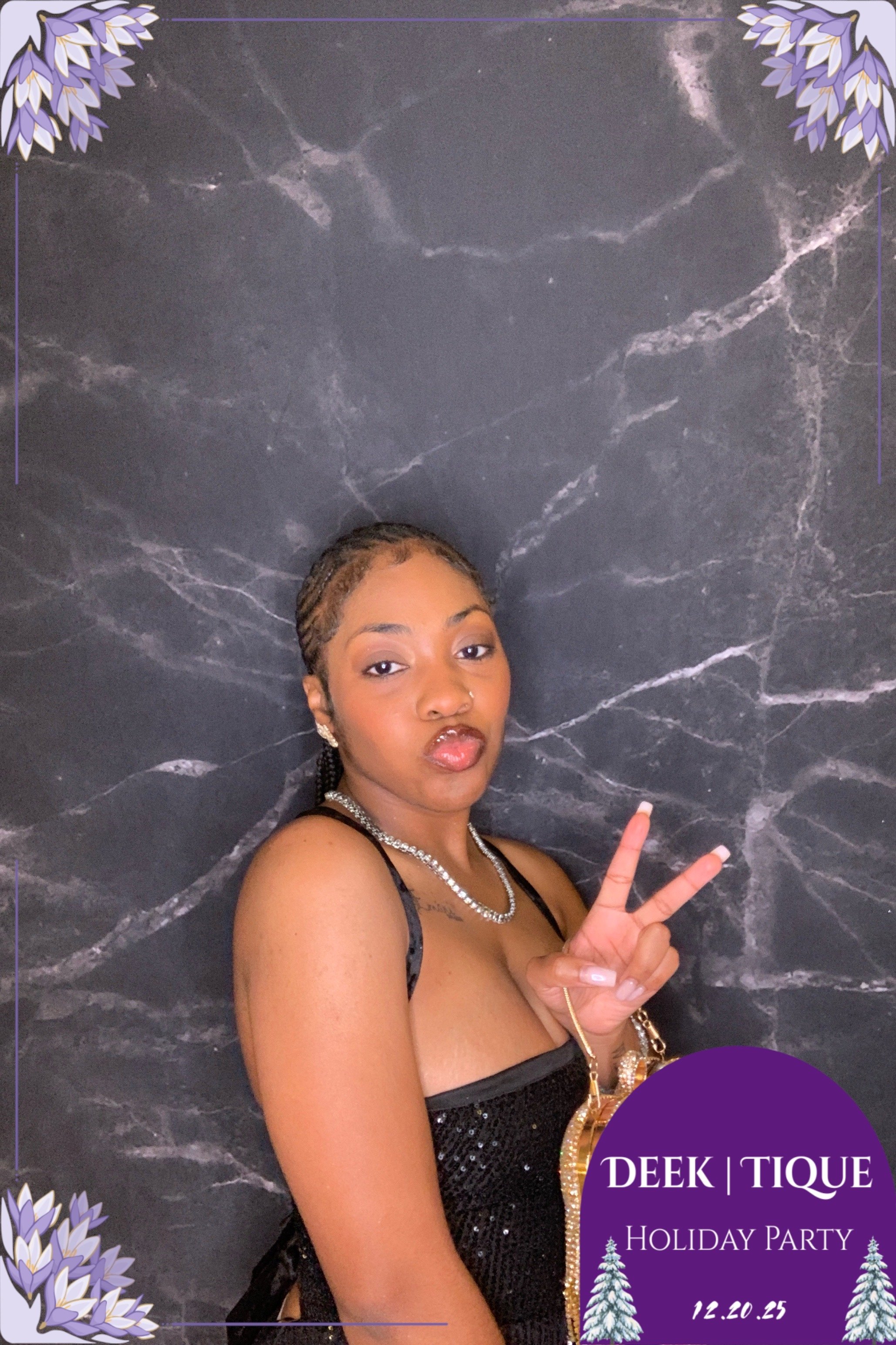 A woman with braided hair taking a selfie in front of a dark marble wall. She is making a peace sign with her right hand, wearing a black, sparkling dress, and accessorized with a necklace and earrings. The photo has a purple-themed Christmas border 