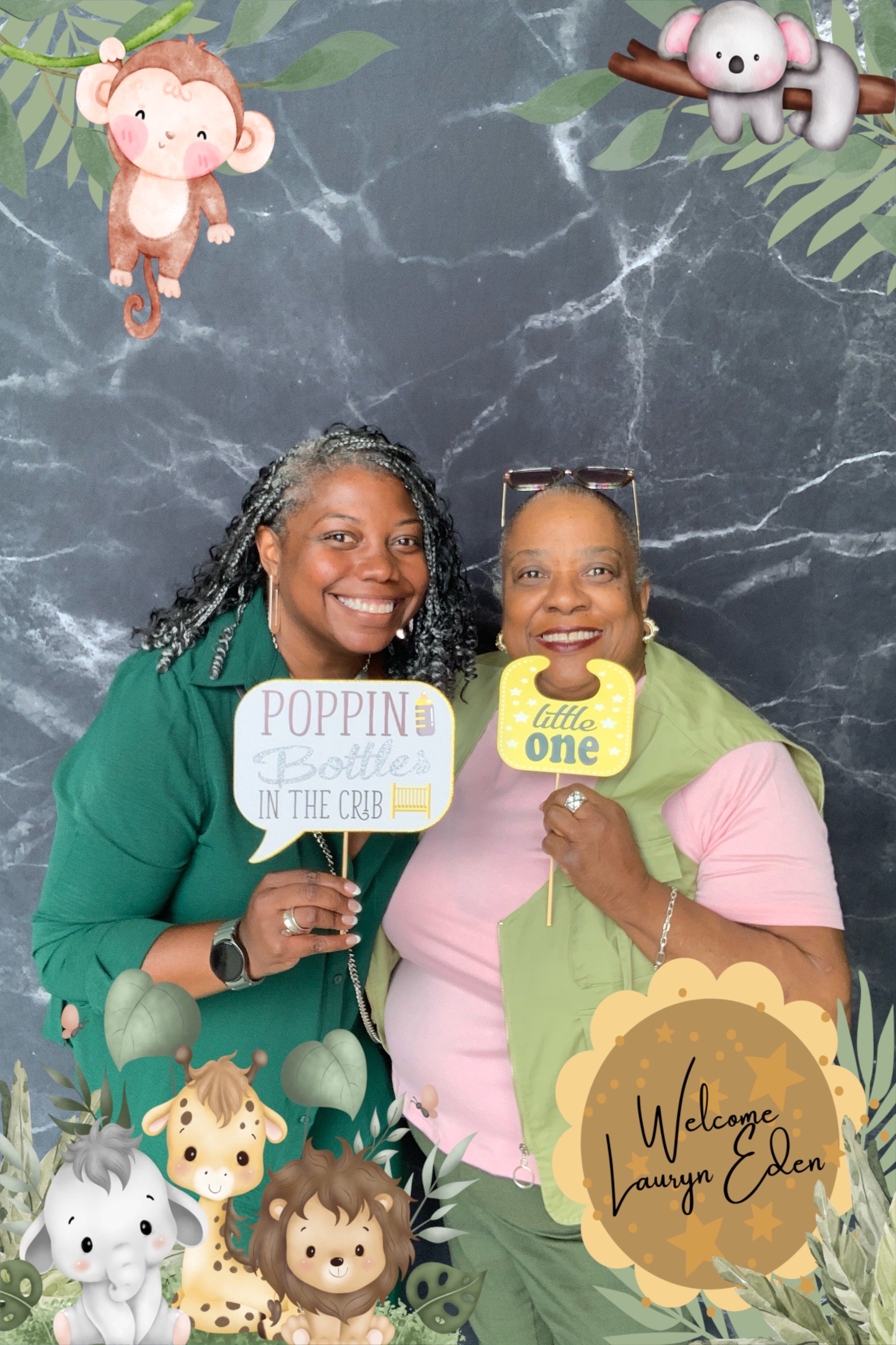 Two women smiling and holding celebration signs, one reading 'POPPIN Bottles IN THE CRIB' and the other 'little ONE'. They are standing in front of a decorative background with cartoon jungle animals and foliage, with a welcome message for Laurnyn Ed