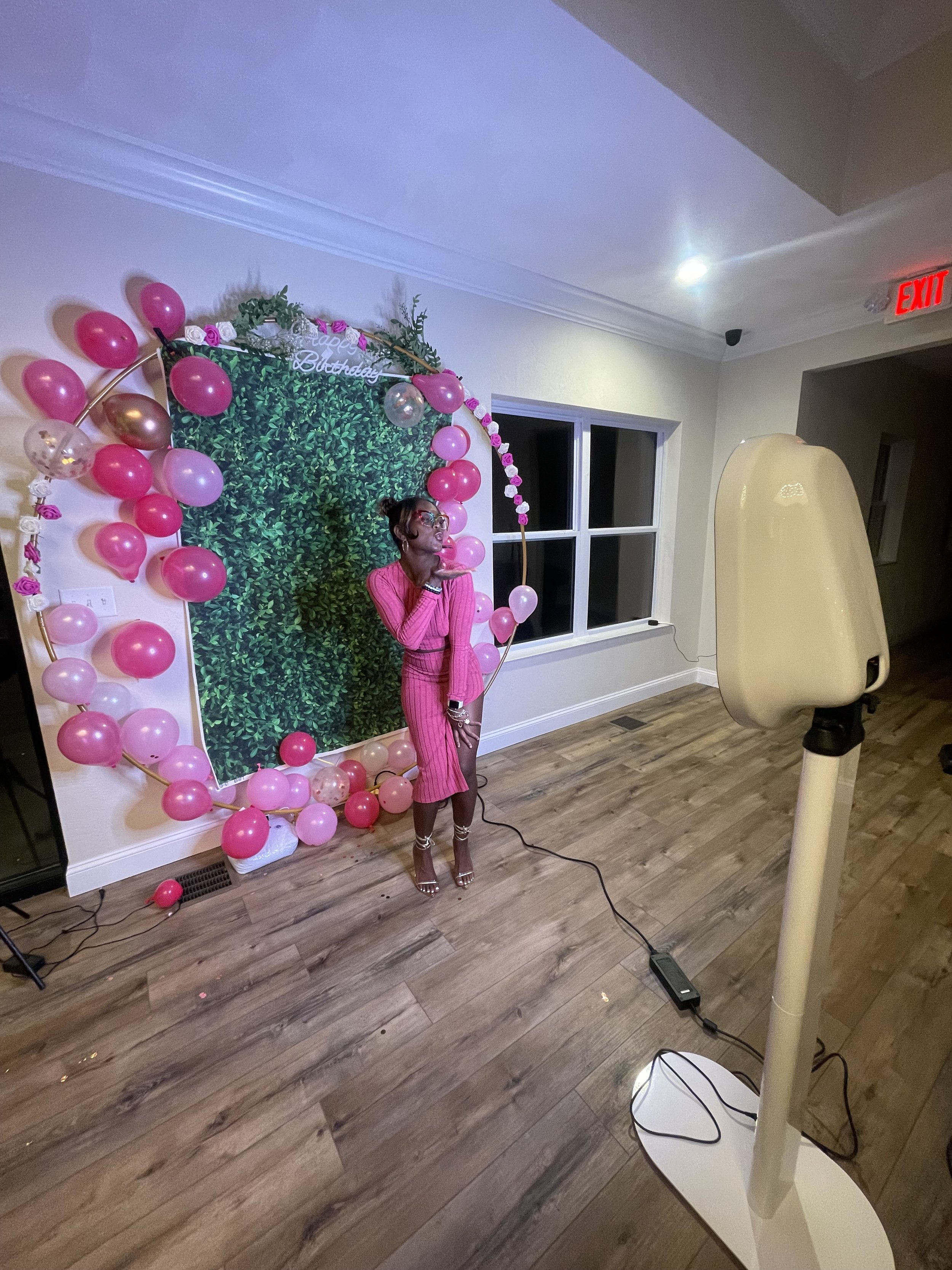A woman in a pink striped outfit stands in front of a green leafy backdrop decorated with pink, purple, and clear balloons. She is posing with one hand near her face. To her right is a large, white photobooth or lighting device, and the room has wood flooring and a window behind her.