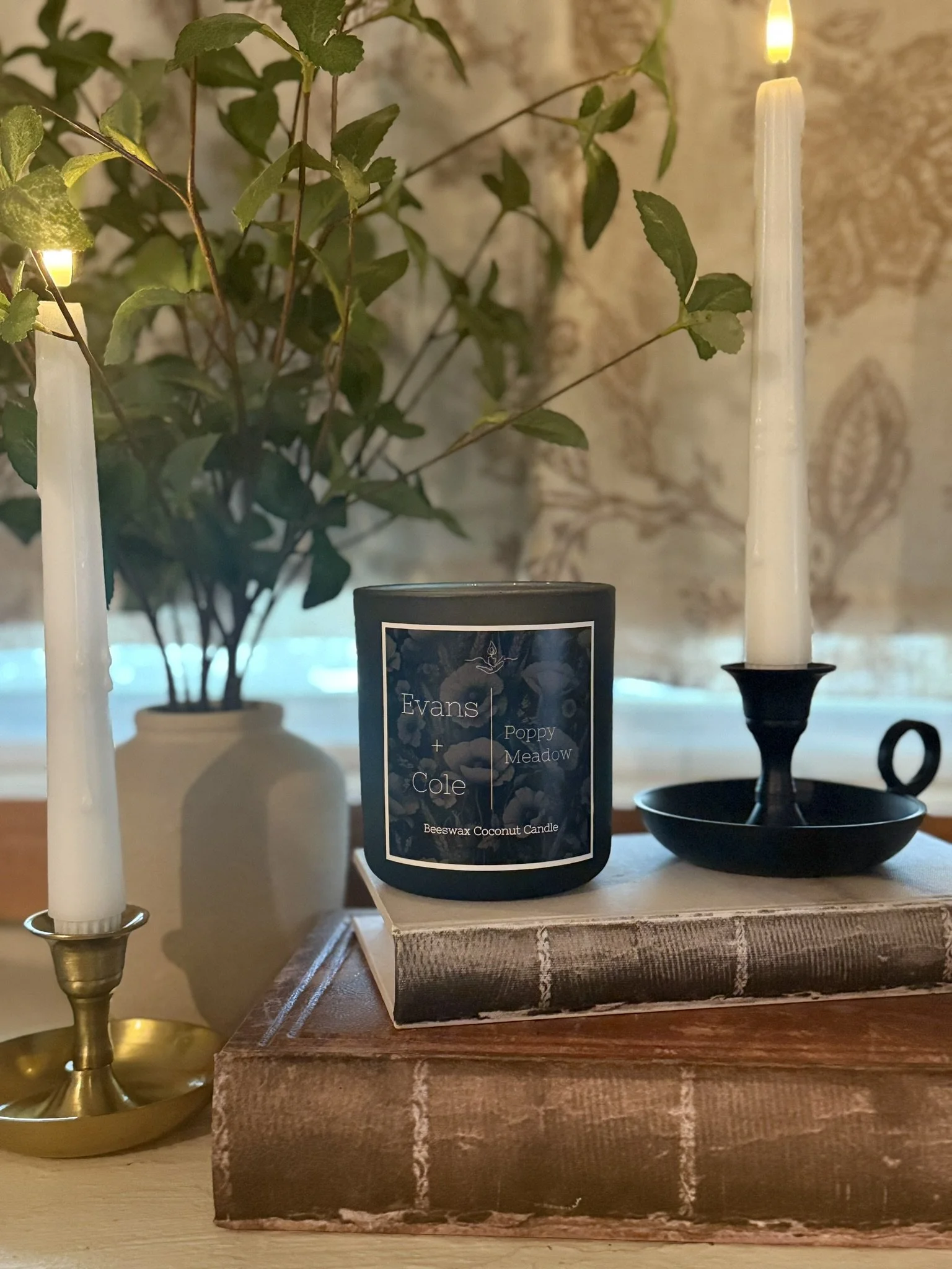 A lit beeswax coconut candle labeled 'Evans + Cole Poppy Meadow' sits on two stacked vintage books, with a candlestick holder and candle on each side, and a large green leafy plant in the background.
