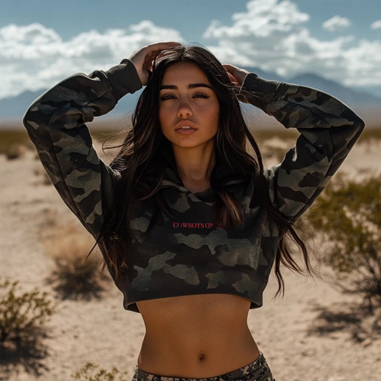 Front view of camo Cowboys Only crop-top hoodie with red text, modeled in rugged desert terrain