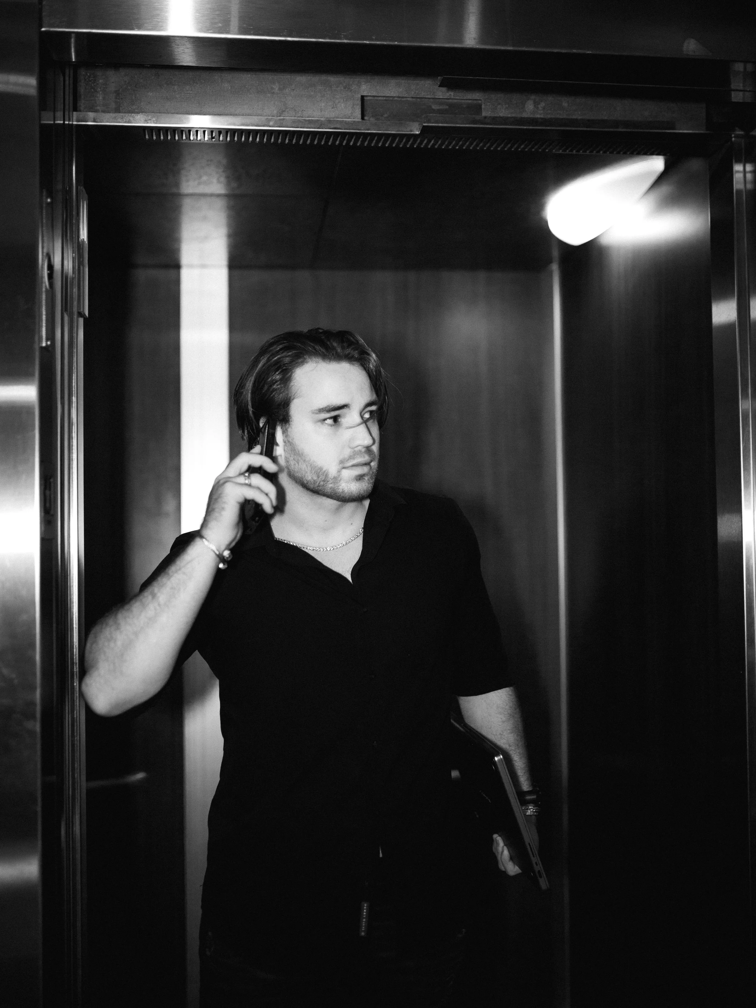 Man in black shirt holding a laptop and phone in an elevator, black and white photo.