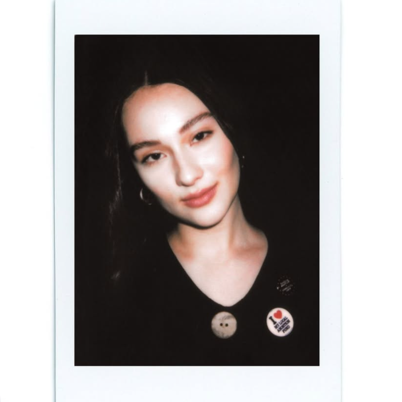 Lola Tung soft smiles in a polaroid by Vincenzo Dimini, wearing two NYAAF pins. One reads "I <3 My Local Abortion Fund," the other says "Funding Abortion = Community Care"