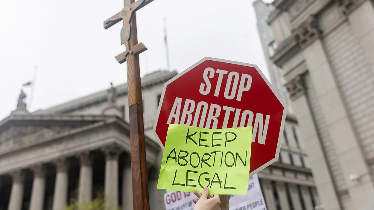 A stop sign says "Stop Abortion." In front of it, a protester holds a sign: "Keep Abortion Legal."
