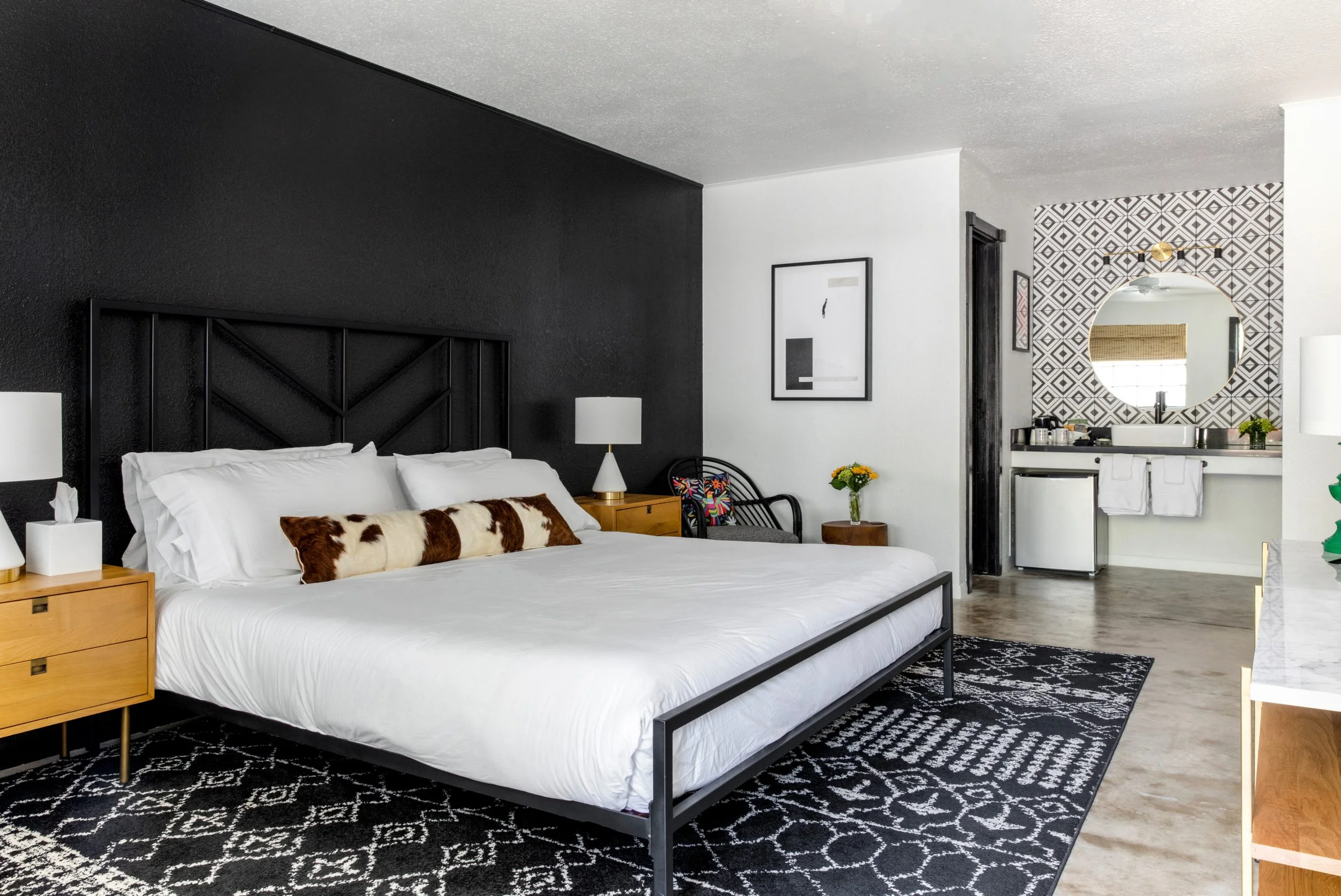 Modern bedroom with black accent wall, white bedding, decorative pillows, wooden nightstands with lamps, patterned rug, chair, wall art, and vanity with round mirror.