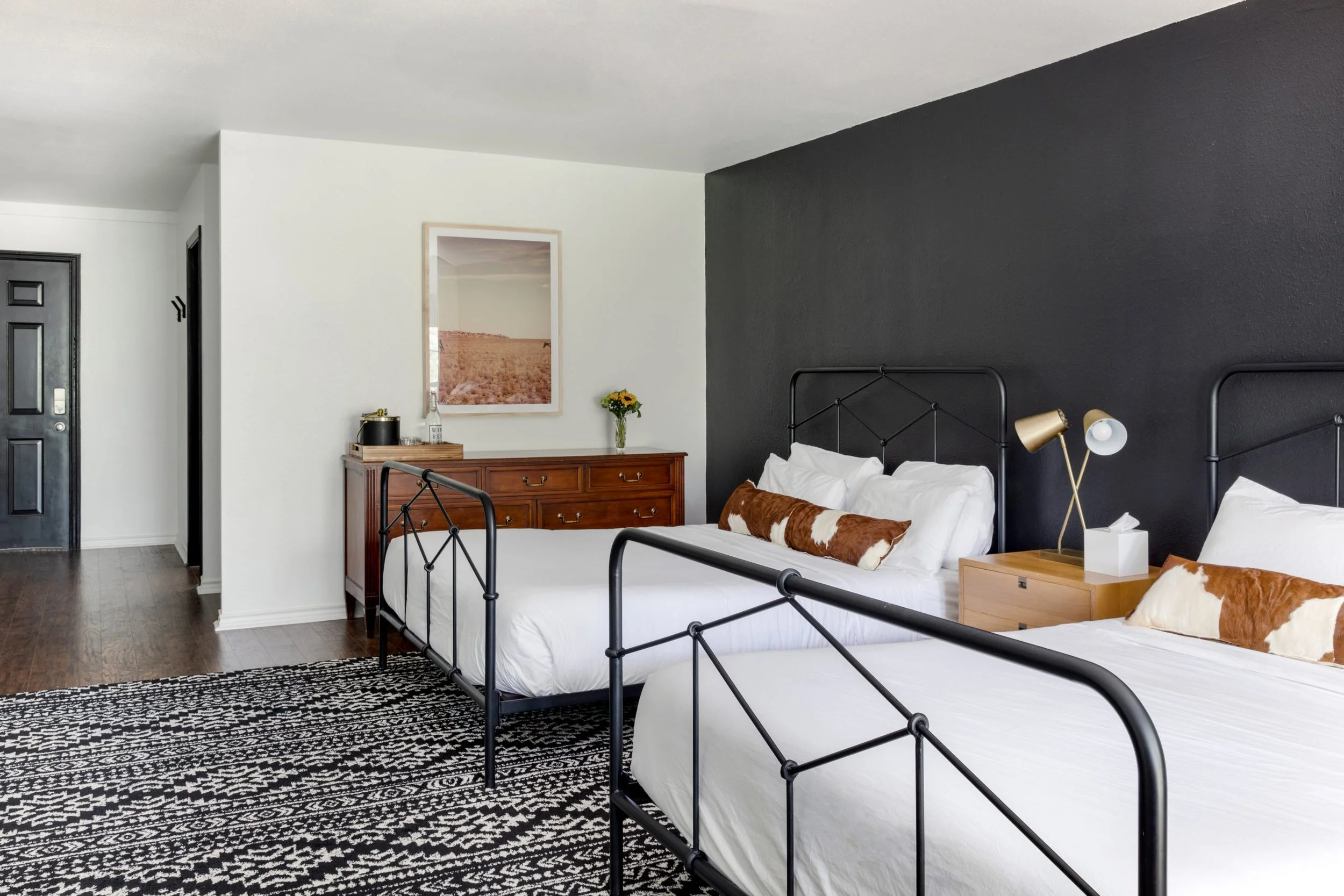 Modern bedroom with two metal-framed beds, white bedding, and cowhide pillows. A wooden dresser with a vase of flowers and a photo frame is against a white wall. The opposite wall is painted black. A patterned rug covers the floor.