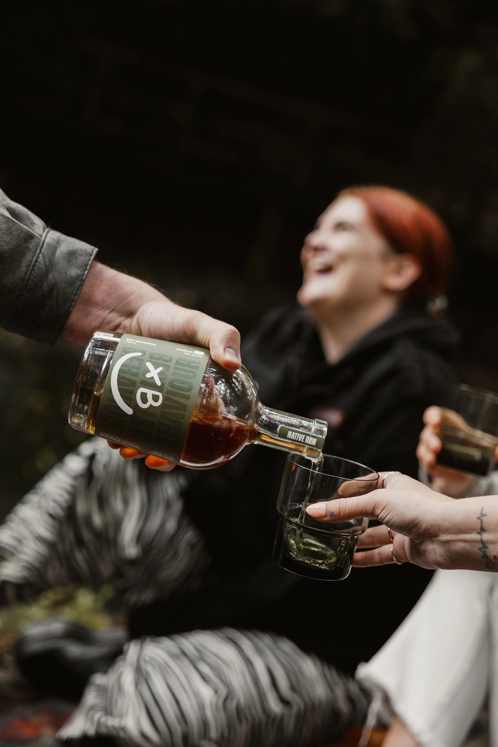 People exchanging drinks, with focus on a hand pouring a beverage from a bottle labeled 'Native Rum' into a glass, and a woman with red hair smiling in the background.