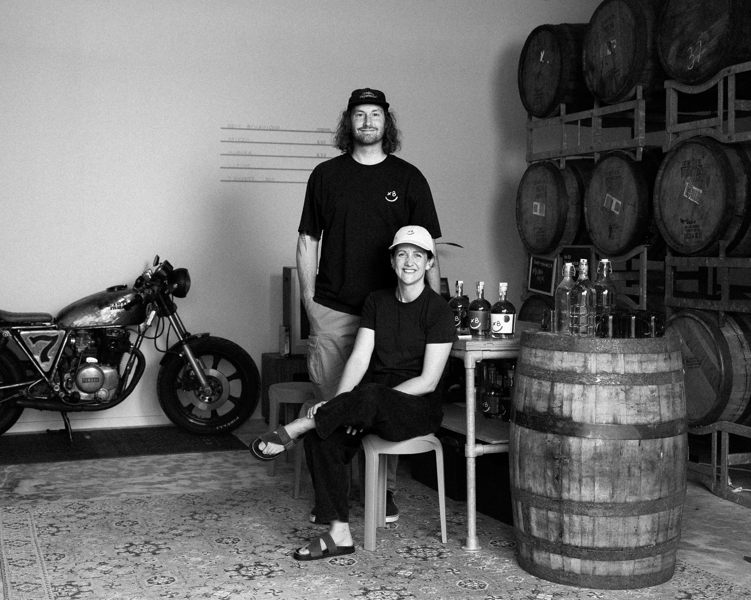 Black and white photo of two smiling people, one standing and one seated, in a room with wine barrels and a motorcycle. The man is tall with curly hair, wearing a black cap and black shirt. The woman, sitting, is wearing a cap and dark clothing. There are bottles on the table and large wine barrels stacked on a rack.