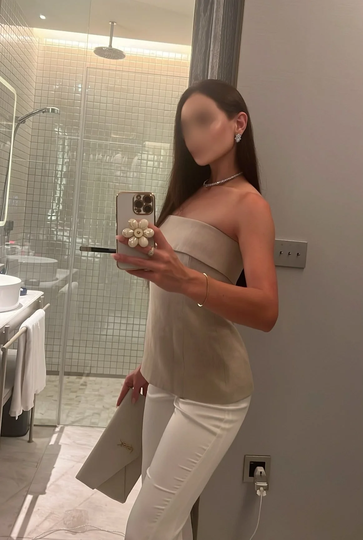 Woman taking a mirror selfie in a bathroom, wearing a strapless beige top, white pants, and jewelry, with a shower and bathroom amenities in the background.