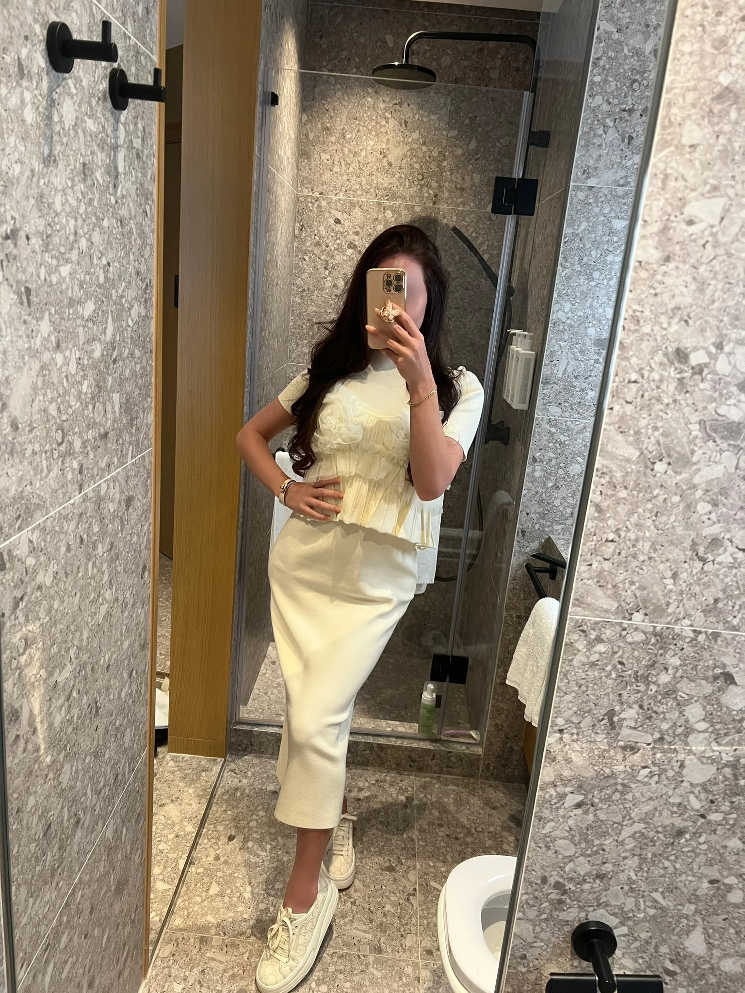 A woman taking a mirror selfie in a bathroom, wearing a cream-colored outfit with sneakers, in front of a glass shower enclosure with grey textured tiles.