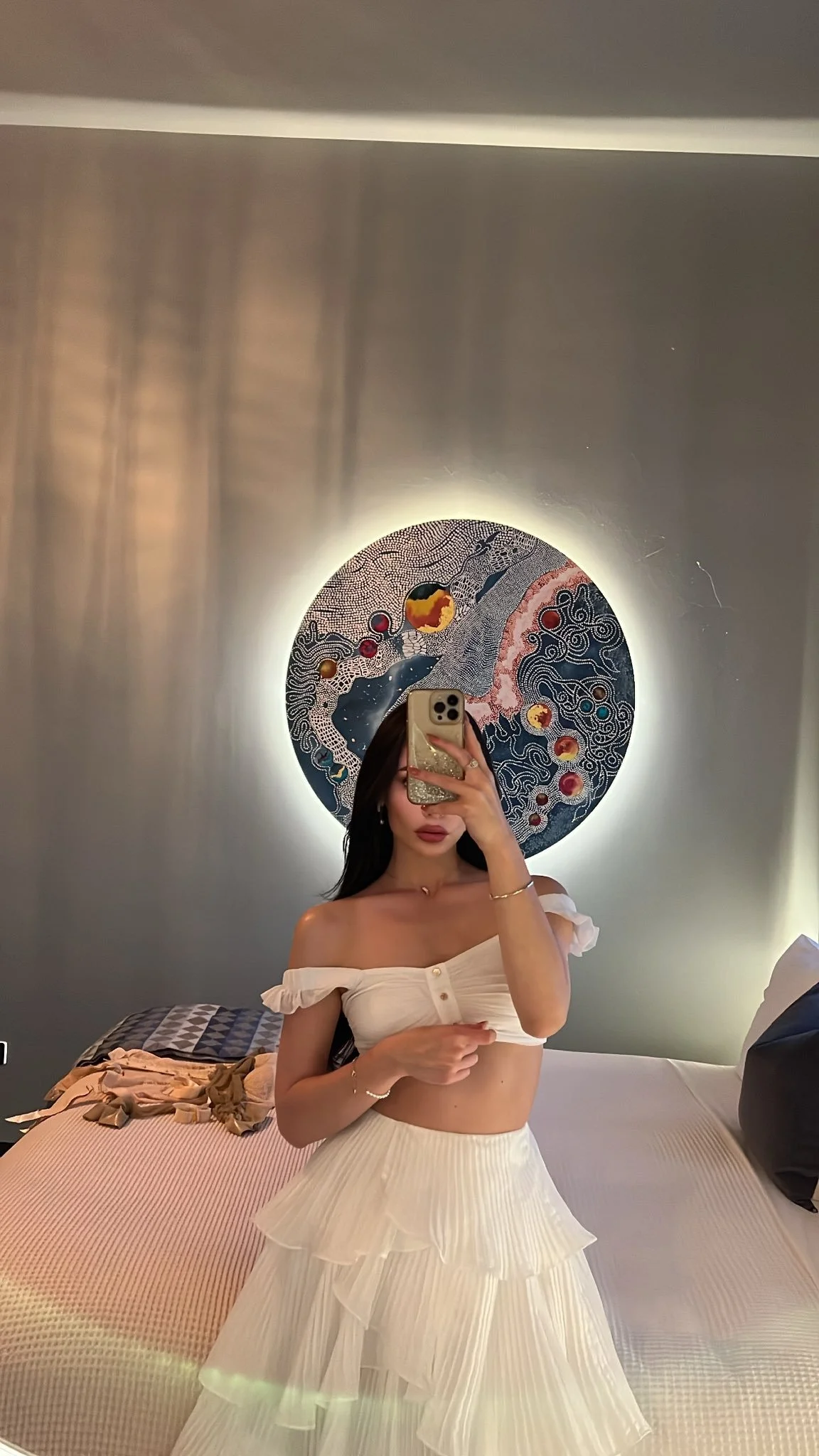 A woman taking a mirror selfie in a hotel room, wearing an off-the-shoulder white crop top and a white tiered skirt. Behind her is a circular artwork with an abstract design, illuminated from behind.