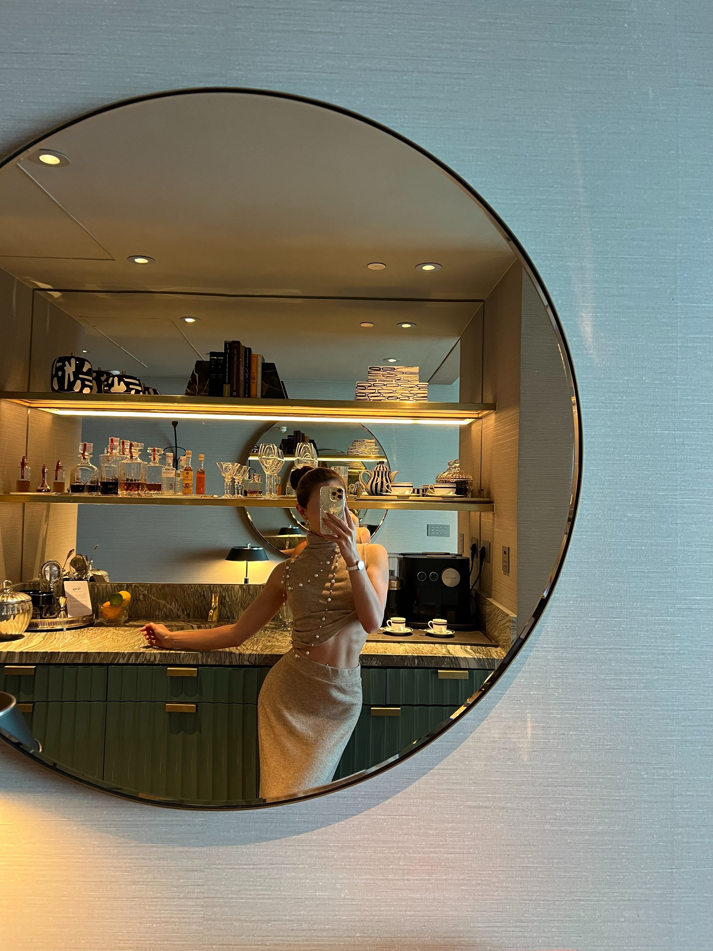A woman taking a mirror selfie in a modern kitchen with a round mirror, decorated with a beige top and matching skirt, her hair tied back, holding a phone.