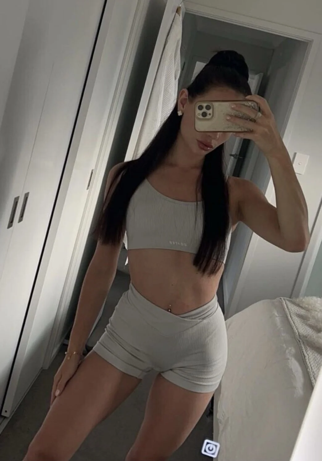 A woman taking a mirror selfie in a bedroom, wearing a light-colored sports bra and matching shorts, with long dark hair and a belly piercing.