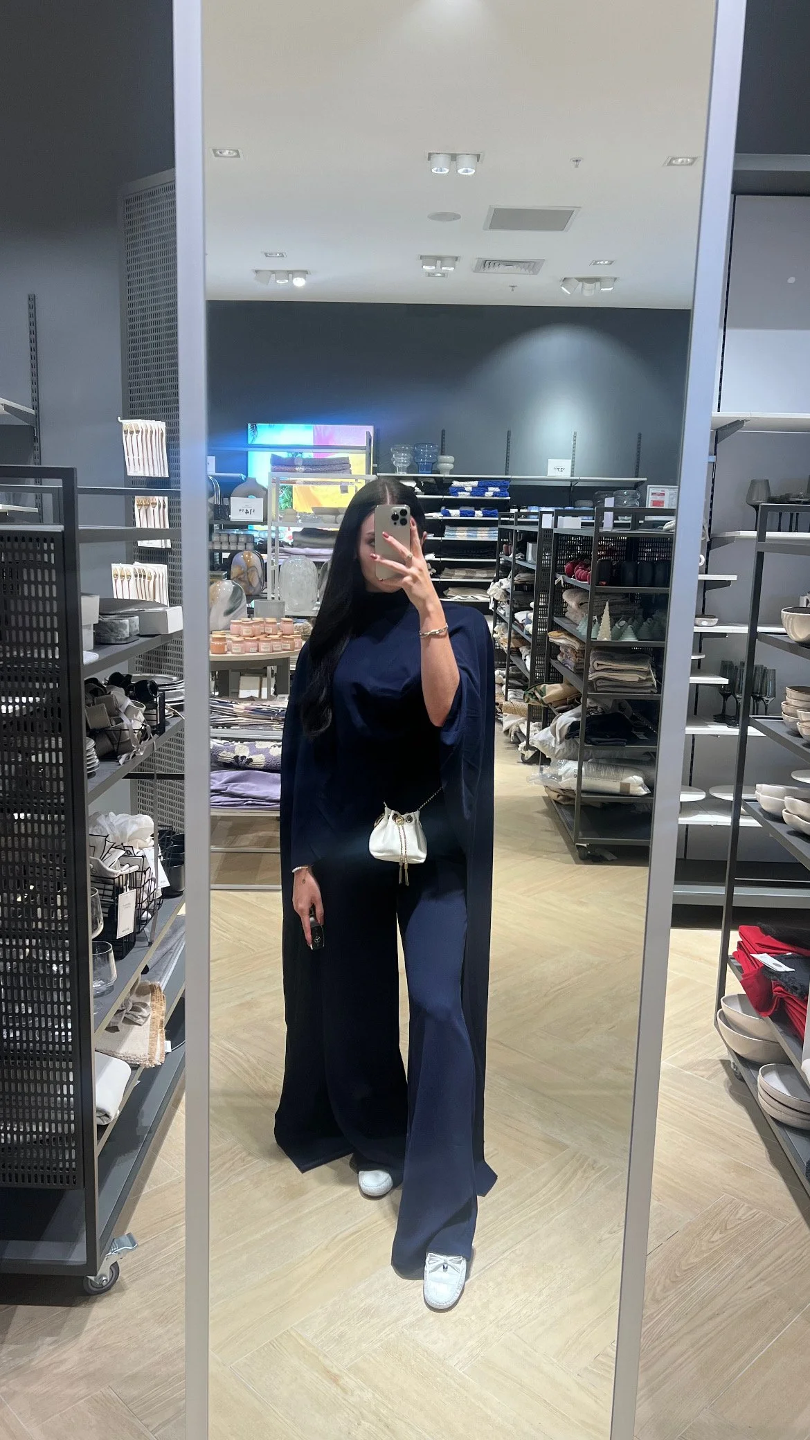 A woman takes a mirror selfie in a home goods store, wearing navy blue wide-leg pants, a matching navy top, white sneakers, and carrying a small white purse.