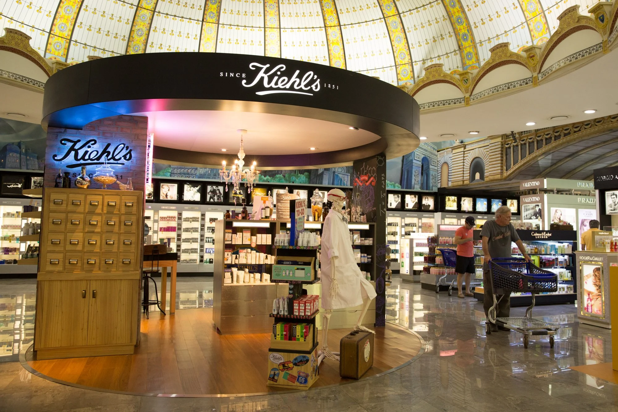 Interior of a store, likely in an airport, with a Kiehl's kiosk at the center. The kiosk has a dark rounded sign with 'Kiehl's' written in white. Inside, there are shelves with skincare products, and a mannequin dressed in white with a vintage suitca