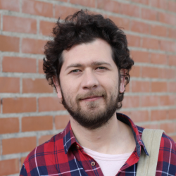 A man with curly dark hair and a beard, wearing a red and blue checkered shirt, standing in front of a brick wall.