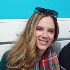 A smiling woman with long blonde hair, wearing sunglasses on her head and a colorful plaid scarf, sitting in front of a blue background.