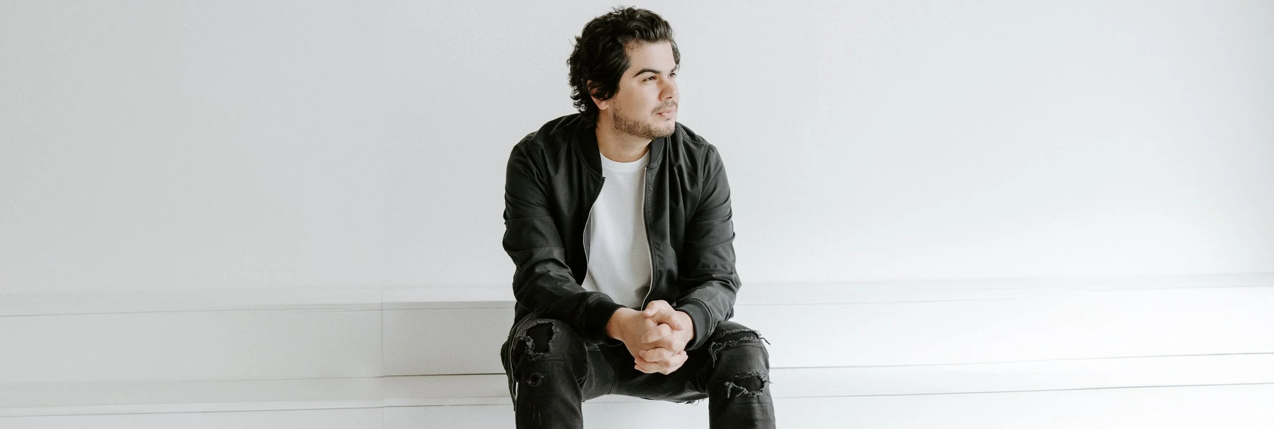 A young man with dark, curly hair and light skin is sitting on the floor against a plain white wall. He is wearing a black jacket over a white shirt and ripped black jeans. He appears to be looking off to the side, lost in thought.